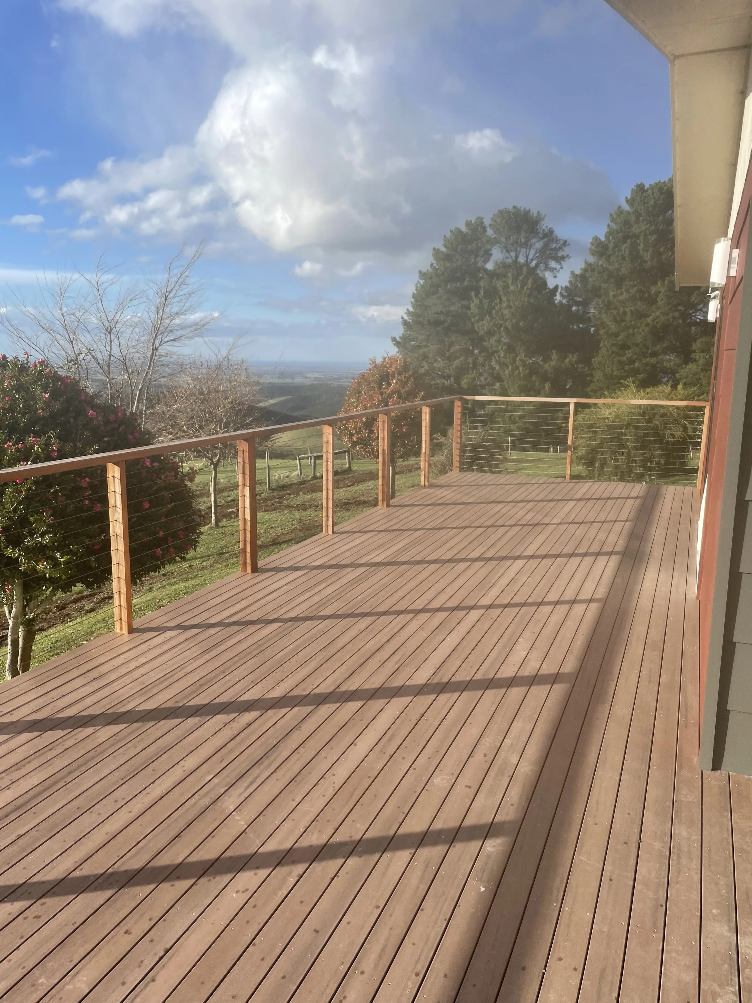 A spacious wooden deck attached to a house, with a metal cable railing, overlooking a scenic landscape of trees, open fields, and a partly cloudy sky.