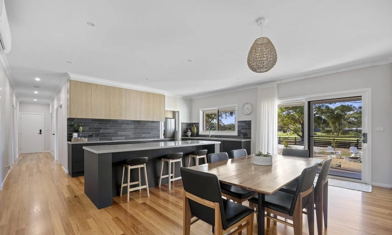 Open kitchen and dining area with wooden floors, black and wood cabinets, gray tile backsplash, and large sliding glass door leading outside to a patio with trees.