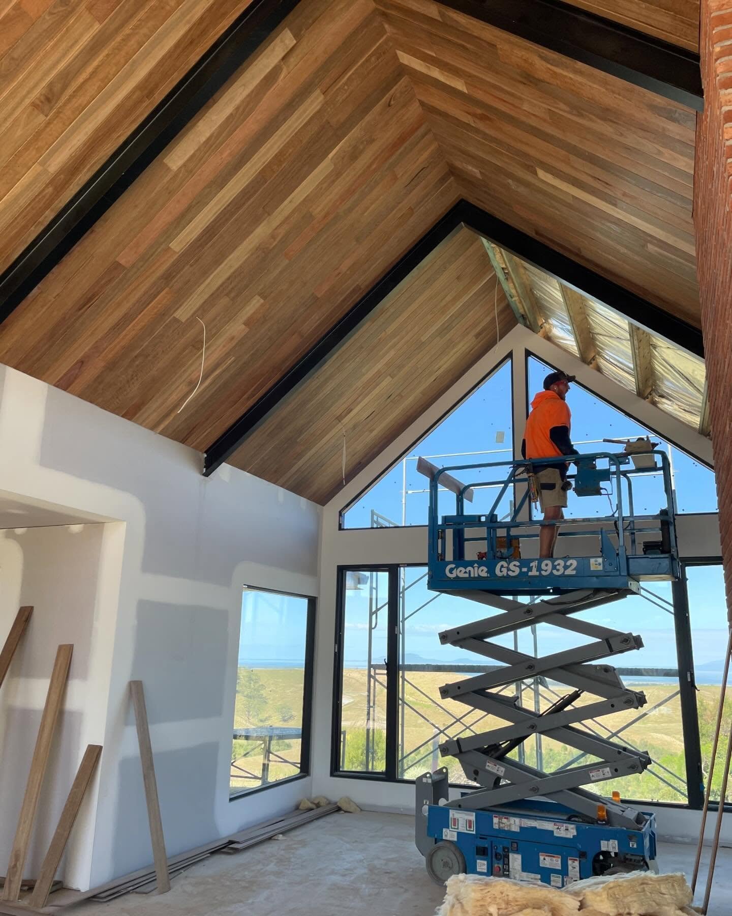 Construction worker on a blue scissor lift installing wooden ceiling panels in a high-ceiling room with large windows, showing blue sky and landscape outside.