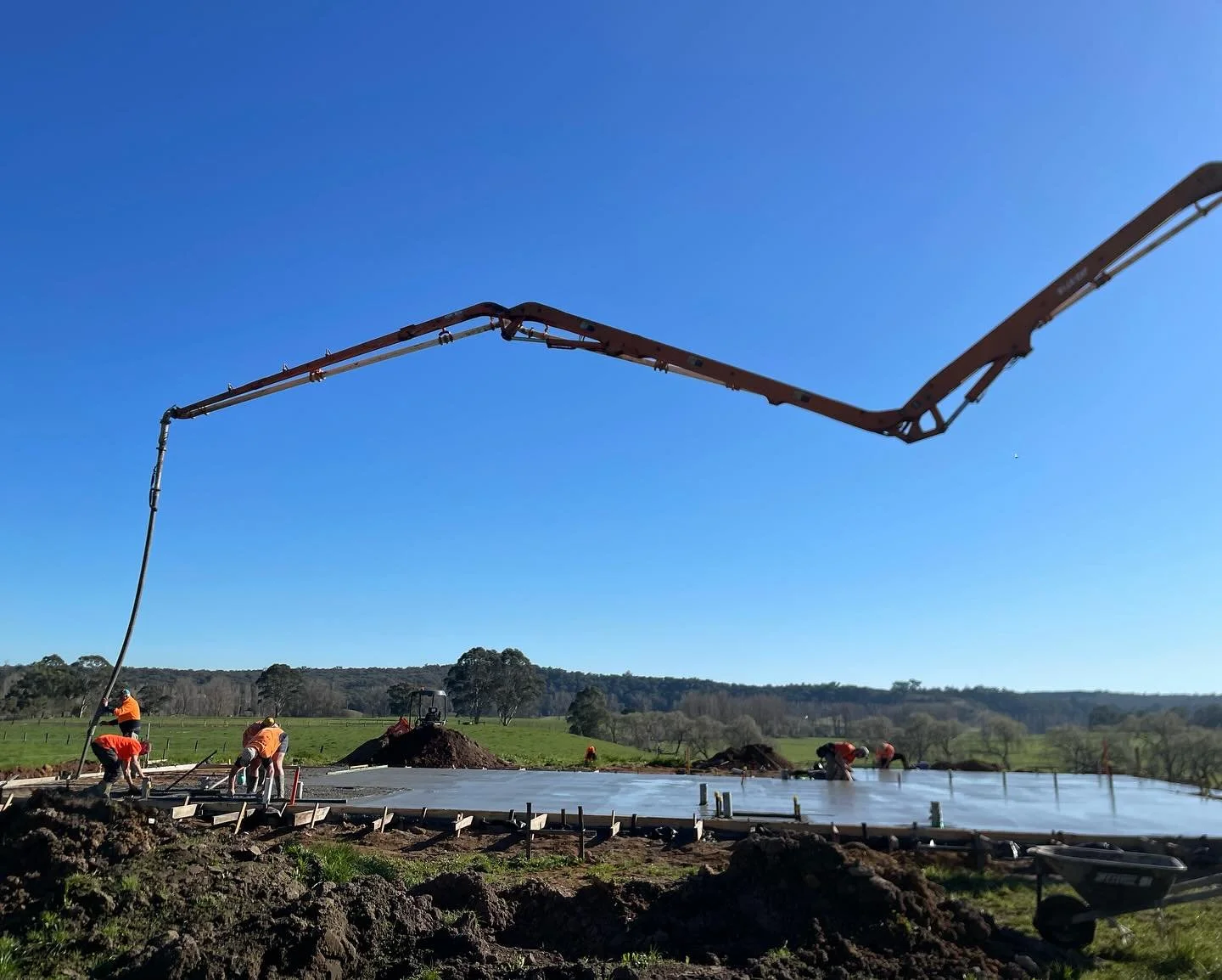 Construction workers are pouring concrete on a building foundation outdoors, with a concrete pump truck pouring the mixture, and a scenic rural landscape in the background under a clear blue sky.