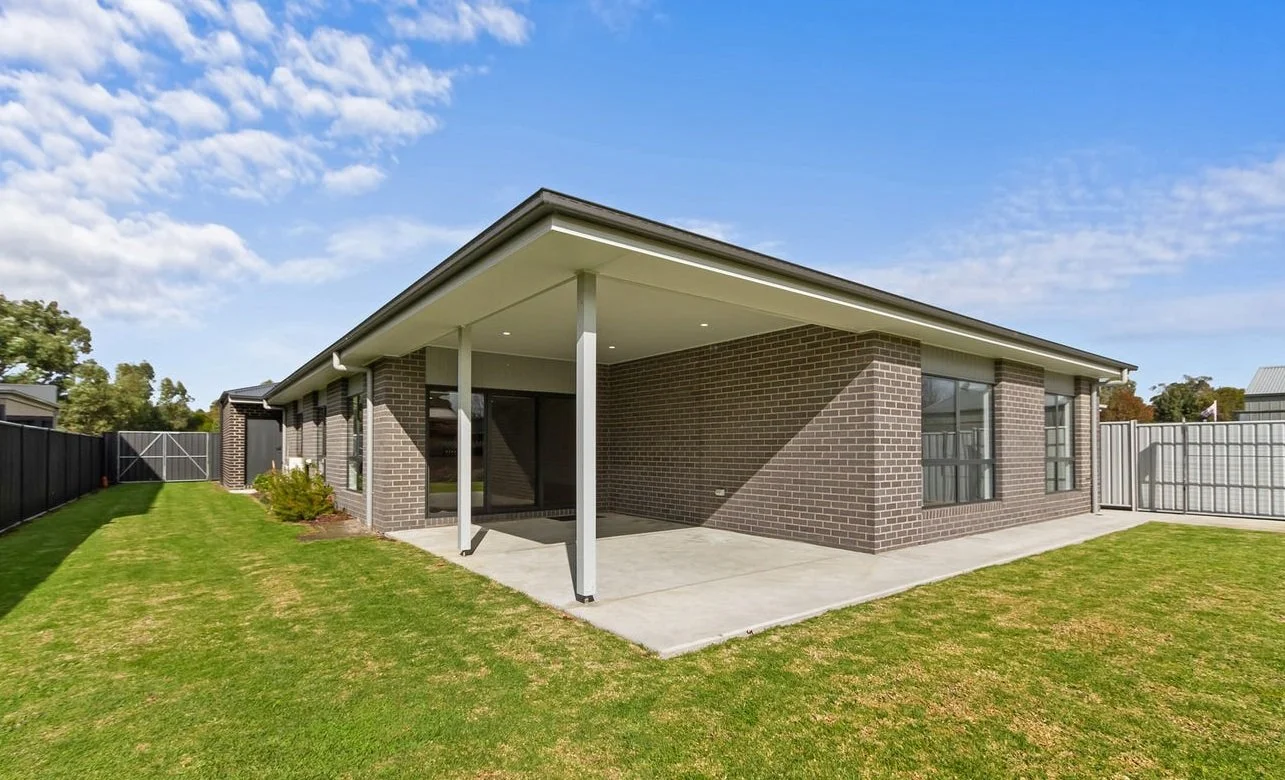 A modern house with brick exterior walls, a covered patio area with a concrete floor, large windows, a sloped roof, and a well-maintained lawn with a black fence surrounding the yard.