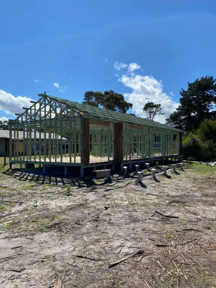 Construction site with a wooden framing of a building under a bright sky, surrounded by trees and open land.