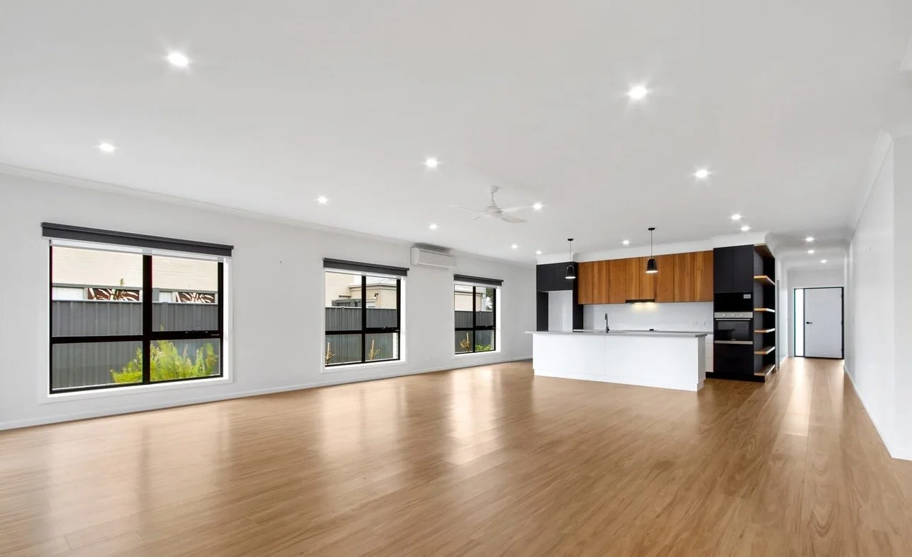 Empty modern living room with large windows, hardwood floors, white walls, and an open kitchen with wooden cabinets, black accents, and pendant lighting.