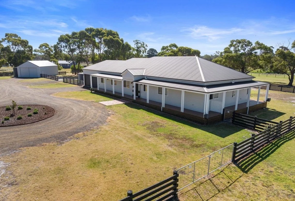 Large white farm house with metal roof and wraparound porch, surrounded by a grassy yard and black wooden fencing, on a sunny day with trees in the background.