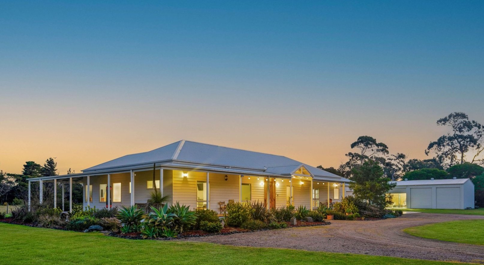 A single-story house with a porch and string lights, situated in a lush yard with landscaping and a gravel driveway on the right, at sunset.