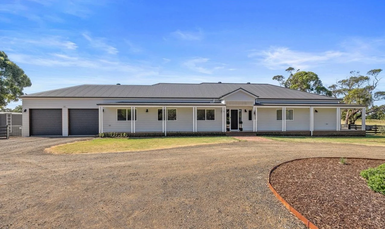 Front view of a modern single-story house with a large gravel driveway, white siding, black garage doors, and a wraparound porch with white columns, under a blue sky with some clouds.