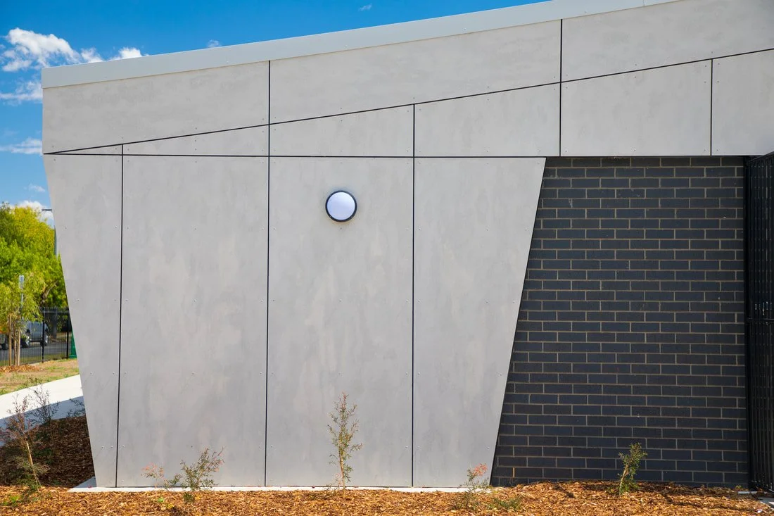 Modern building exterior with light grey concrete panels and dark brick accents, under a blue sky with white clouds.