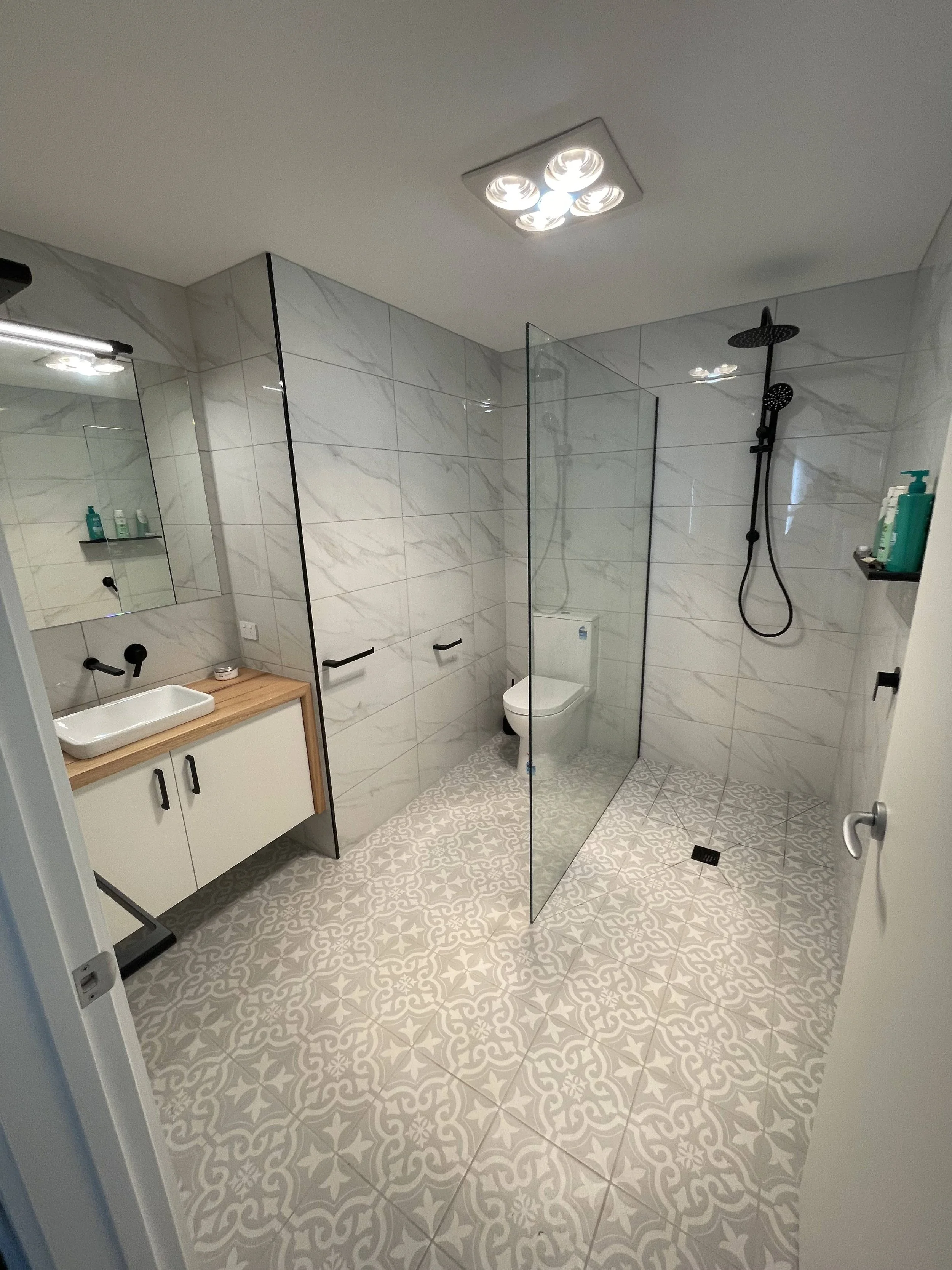 Modern bathroom with patterned tile floor, marble wall tiles, a sink with a wooden countertop, a mirror, a glass shower partition, a ceiling light, a wall-mounted toilet, and a black shower system with a rain shower head.