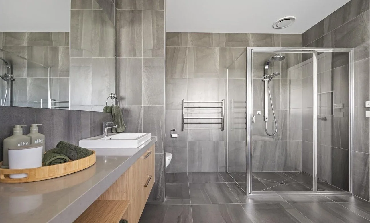 Modern bathroom with gray tiled walls and floor, glass shower enclosure, wall-mounted towel warmer, vanity with sink, mirror, and toiletries.