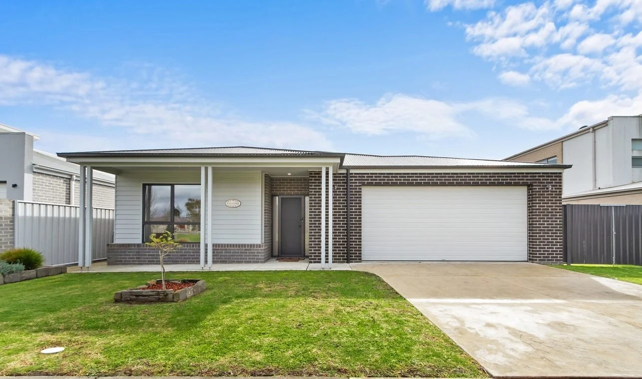 Front view of a modern house with a green lawn, a small tree in a flower bed, a concrete driveway, and a two-car garage under a blue sky with scattered clouds.