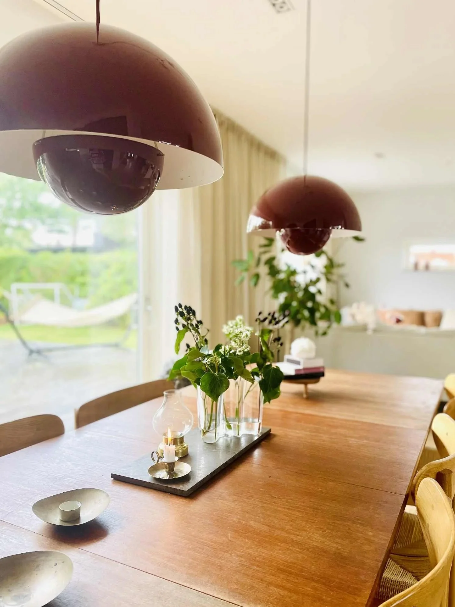 Et langt trebord med en blomsterarrangement i midten, pyntet med lys og dekorative gjenstander, i et rom med store vinduer og gardiner. To hengelamper henger over bordet, og det er lenestoler rundt bordet.