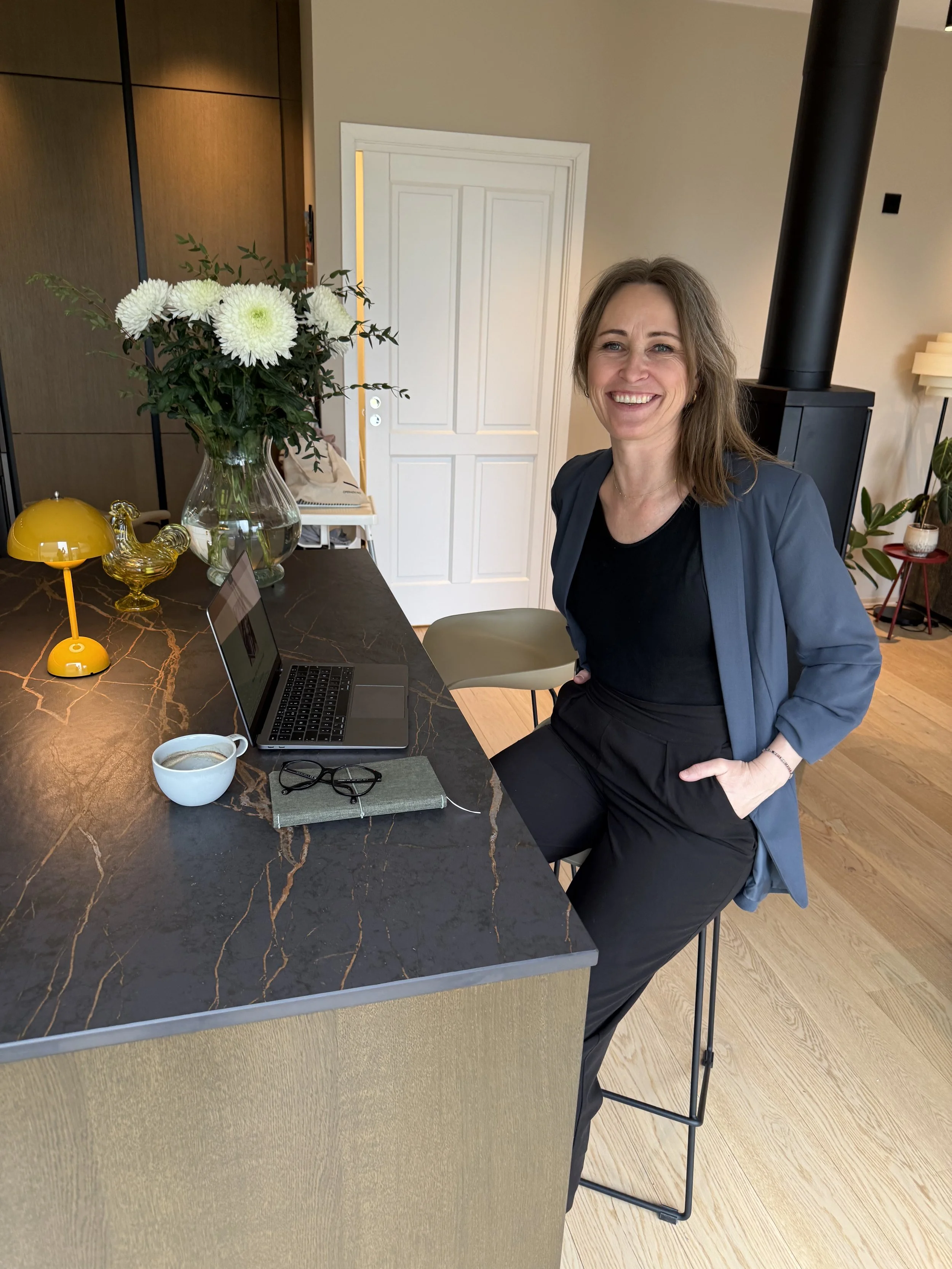En kvinne med brunt hår smiler i et rom, står ved en kjøkkenøy av marmor. Det er en åpen plante og dekorative gjenstander på bordet, inkludert en gul lampe, en gitt, en kopp med kaffe, en bærbar datamaskin og et par briller. I bakgrunnen er en hvit dør, et svart røykrør og innervegger i beige.