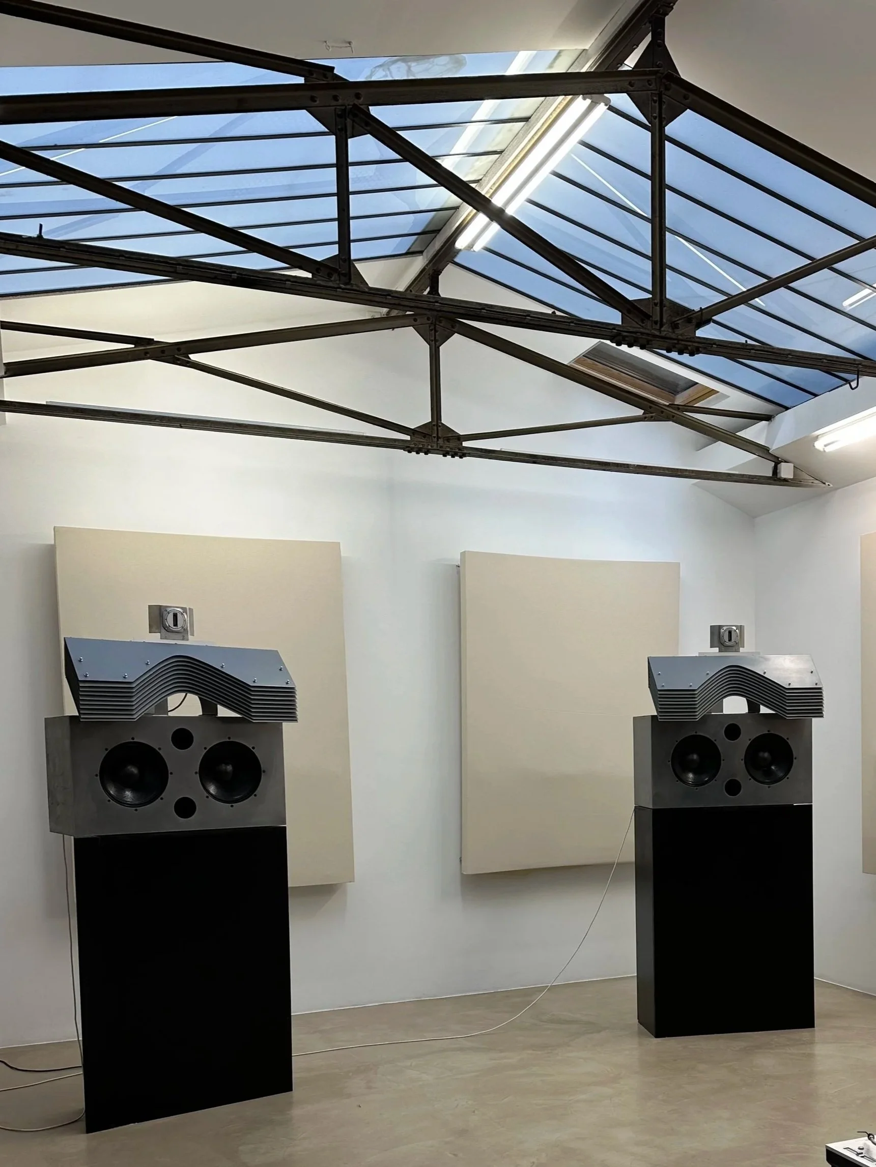 Deux haut-parleurs de grande taille sont placés sur des piédestaux noirs dans une pièce d'exposition. La pièce possède une paroi en arrière avec des panneaux acoustiques, et une toiture en verre permettant de voir le ciel.