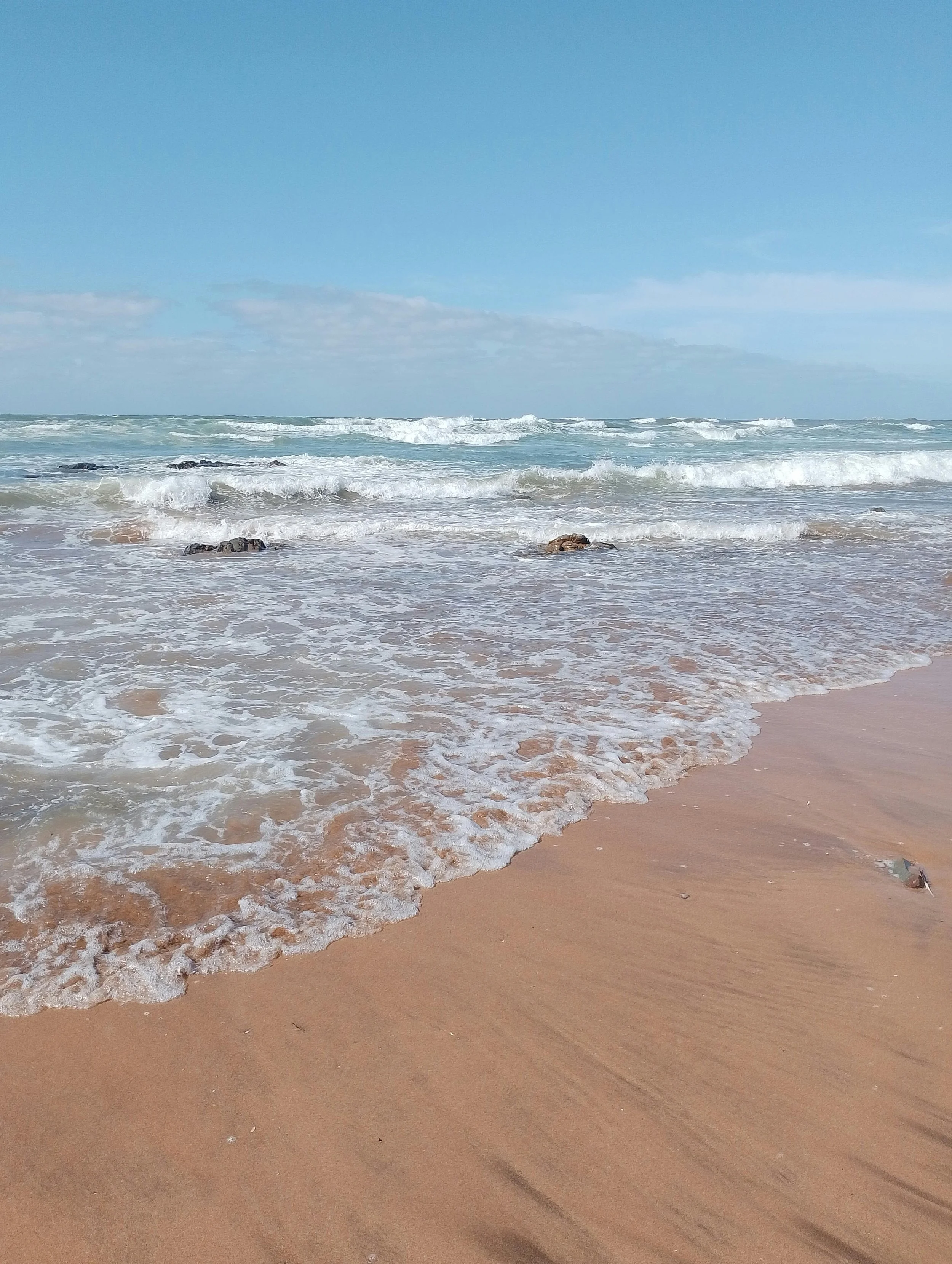 Plage de sable avec vagues, quelques rochers, ciel bleu avec quelques nuages.