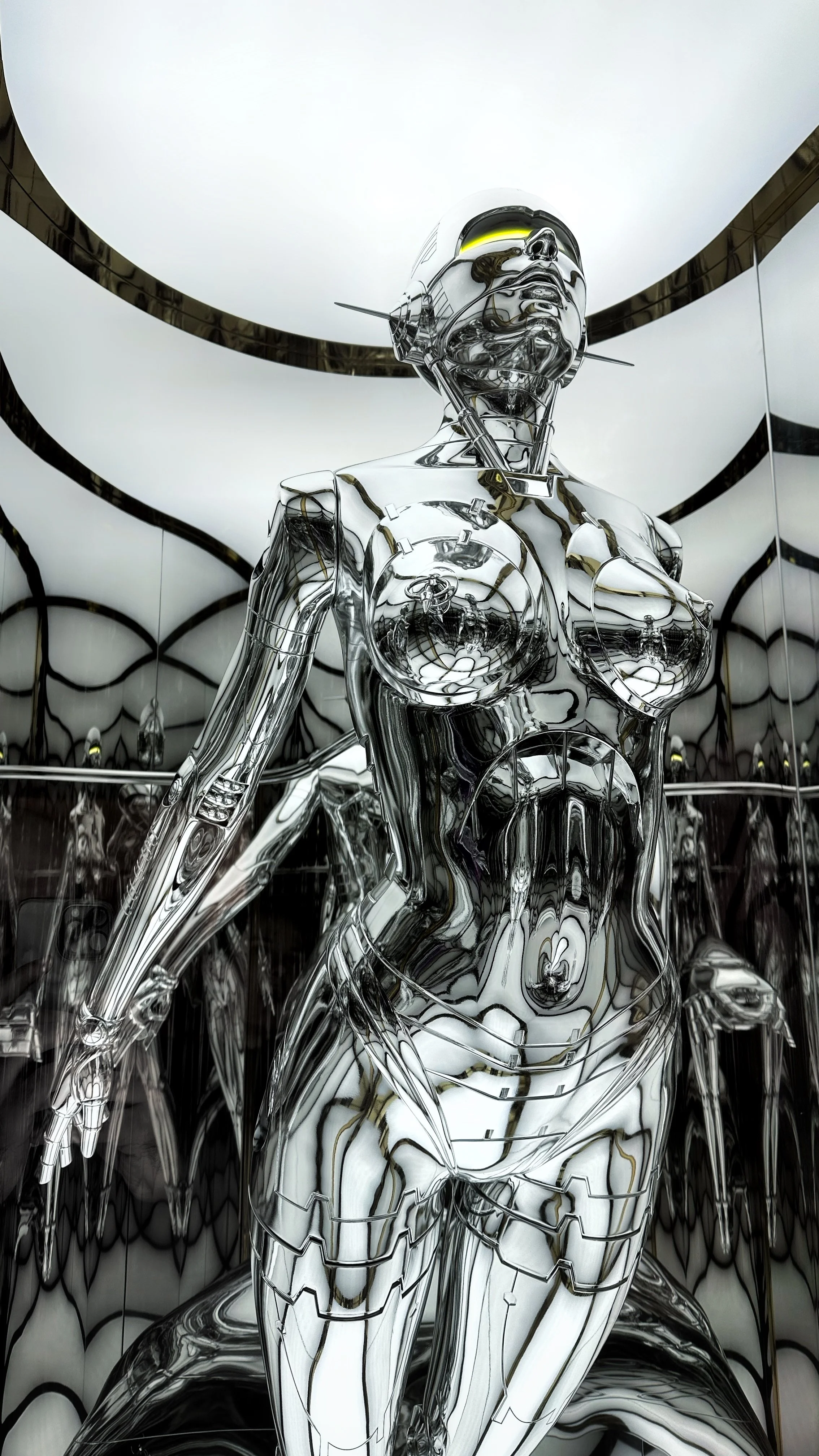 Une sculpture futuriste en métal poli représentant un corps féminin avec un casque et des éléments mécaniques, dans un espace entouré de miroirs.