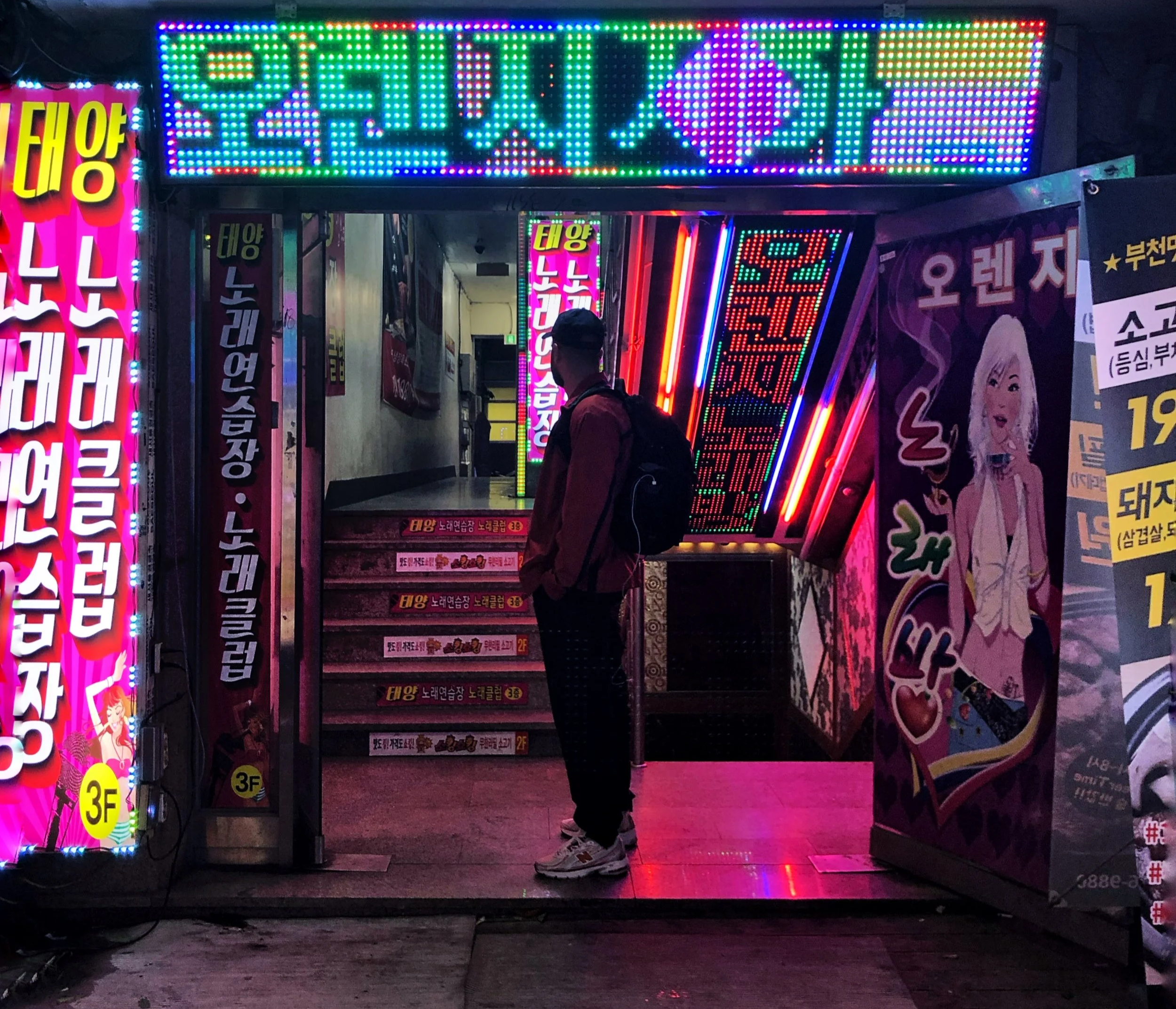 Une personne avec un sac à dos regarde l'entrée illuminée d'un bâtiment décoré de lumières LED multicolores avec des panneaux et affiches en coréen, dans un environnement urbain nocturne.