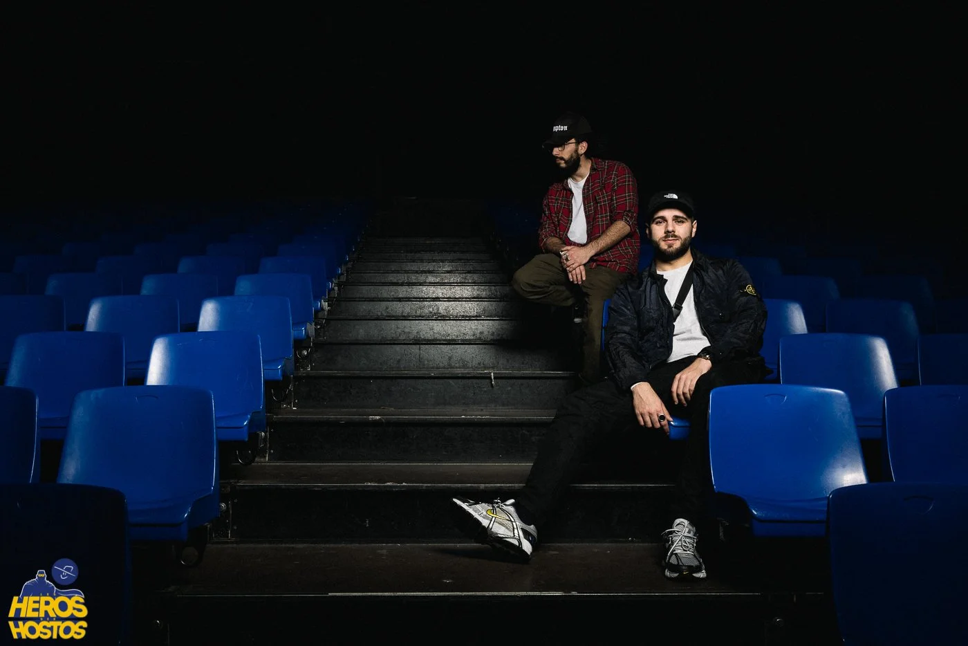 Deux hommes assis sur des gradins bleus dans une salle sombre, l'un en train de se tenir debout, l'autre assis avec une casquette, vêtements décontractés, ambiance sombre et mystérieuse.