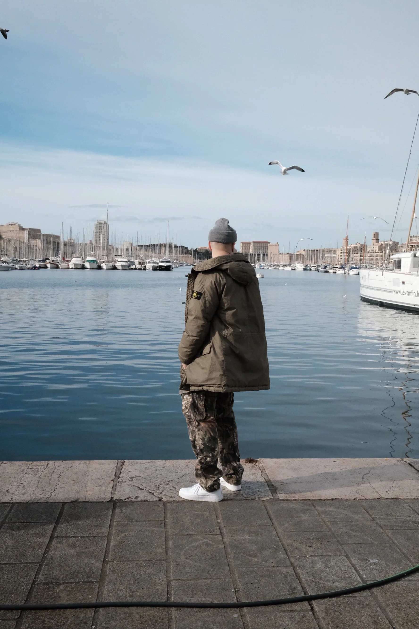 Homme debout au bord d'une marina regardant la mer avec des mouettes volant dans le ciel, en arrière-plan des bateaux amarrés et une ville.