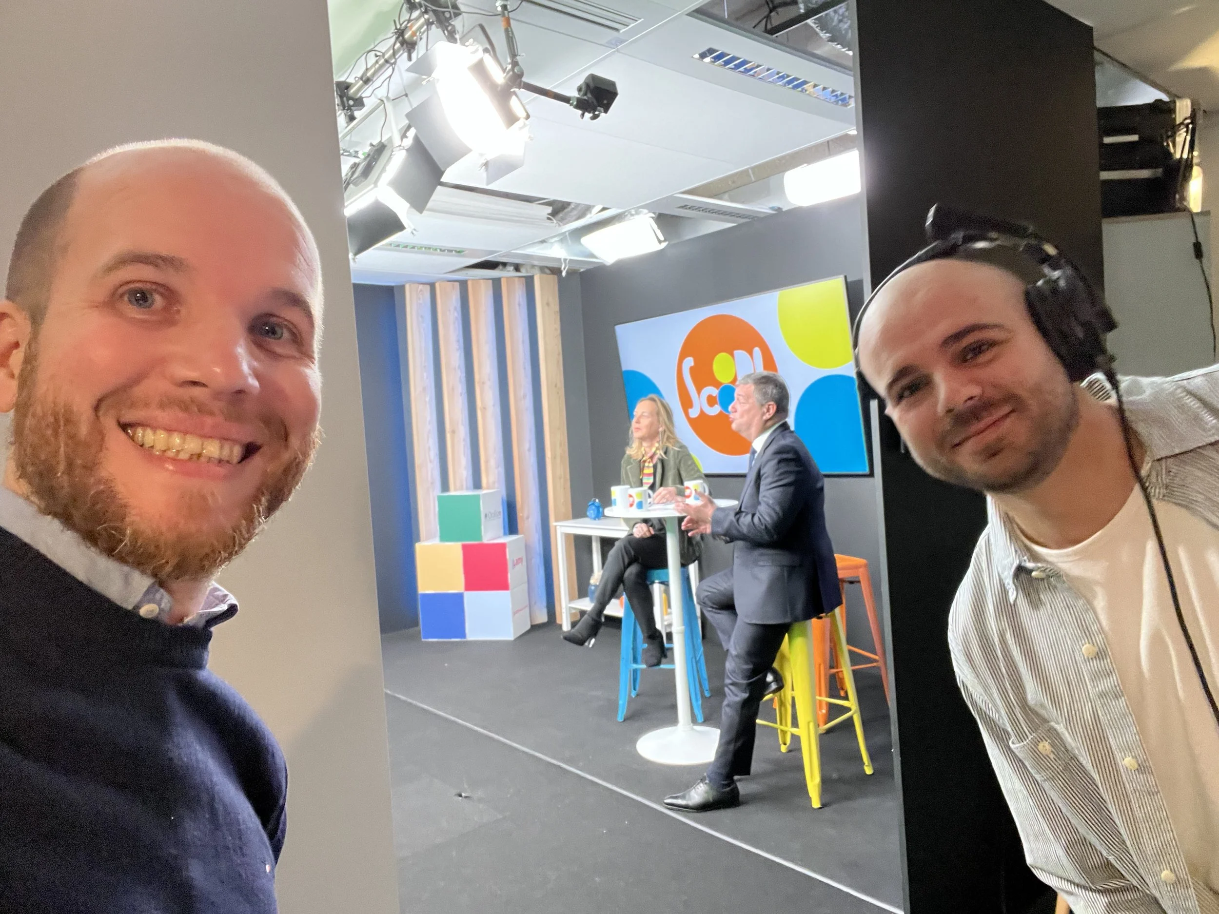 Deux hommes prennent un selfie devant un studio de télévision avec deux personnes assises derrière eux. Les deux personnes à l'arrière sont en pleine émission, un écran derrière elles affiche un logo coloré 'Scoop'.