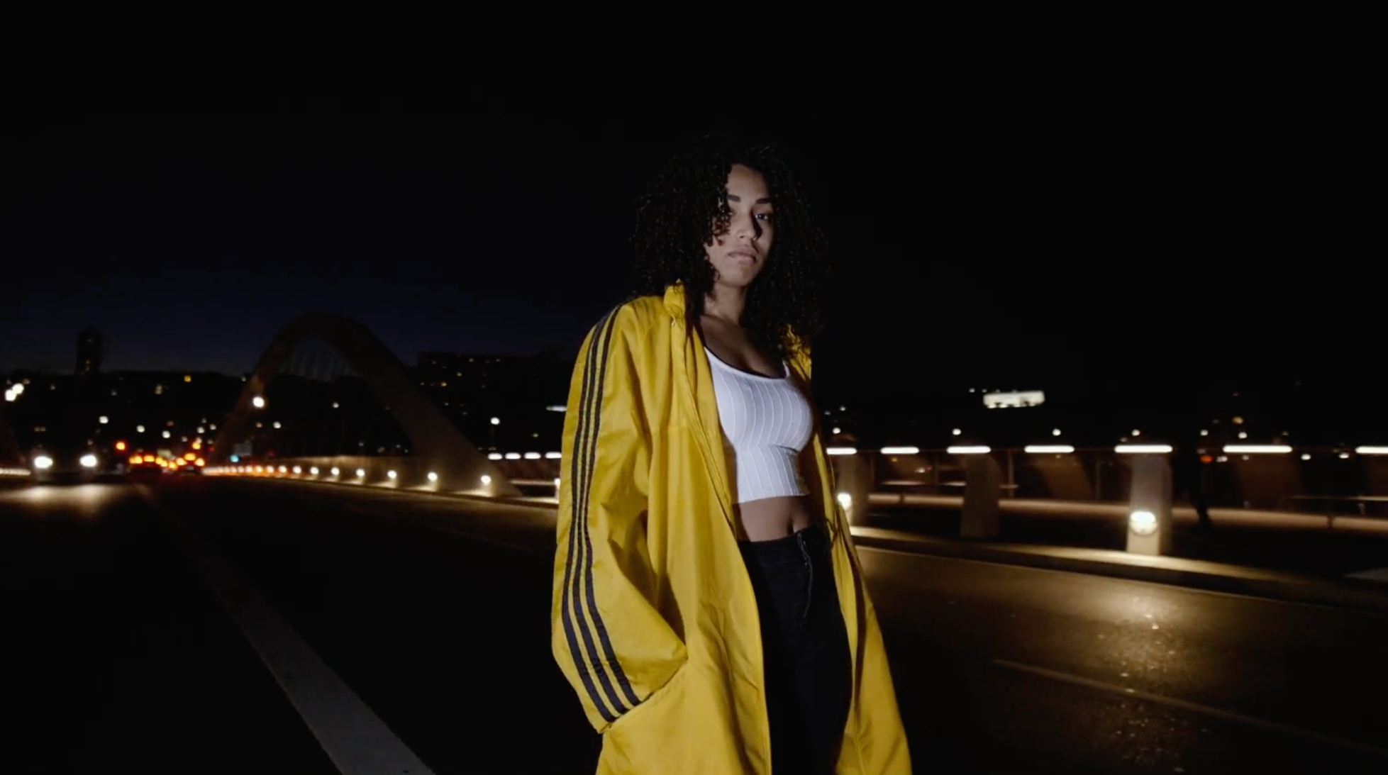 Jeune femme portant une veste jaune et un haut blanc, debout sur un pont la nuit, avec des lumières urbaines en arrière-plan.