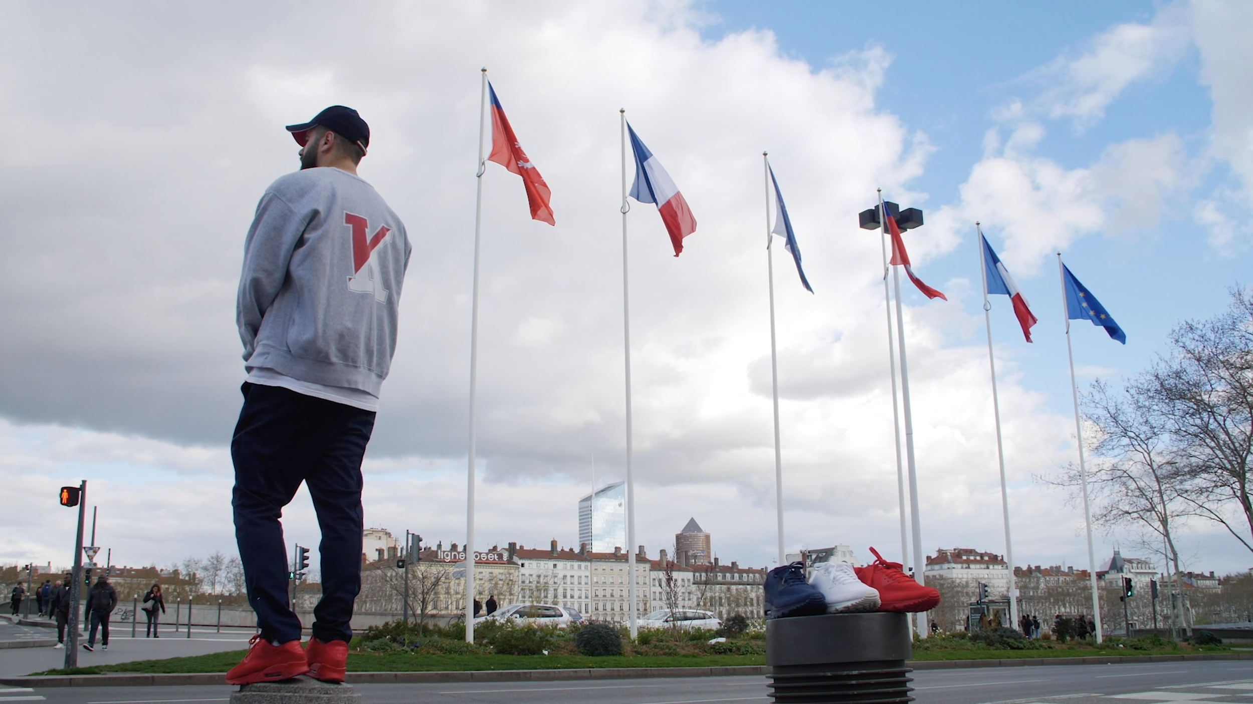 Un homme debout près d'un panneau de drapeaux européens et français, portant une veste grise avec la lettre V sur le dos, dans une ville avec des bâtiments urbains et des nuages dans le ciel.