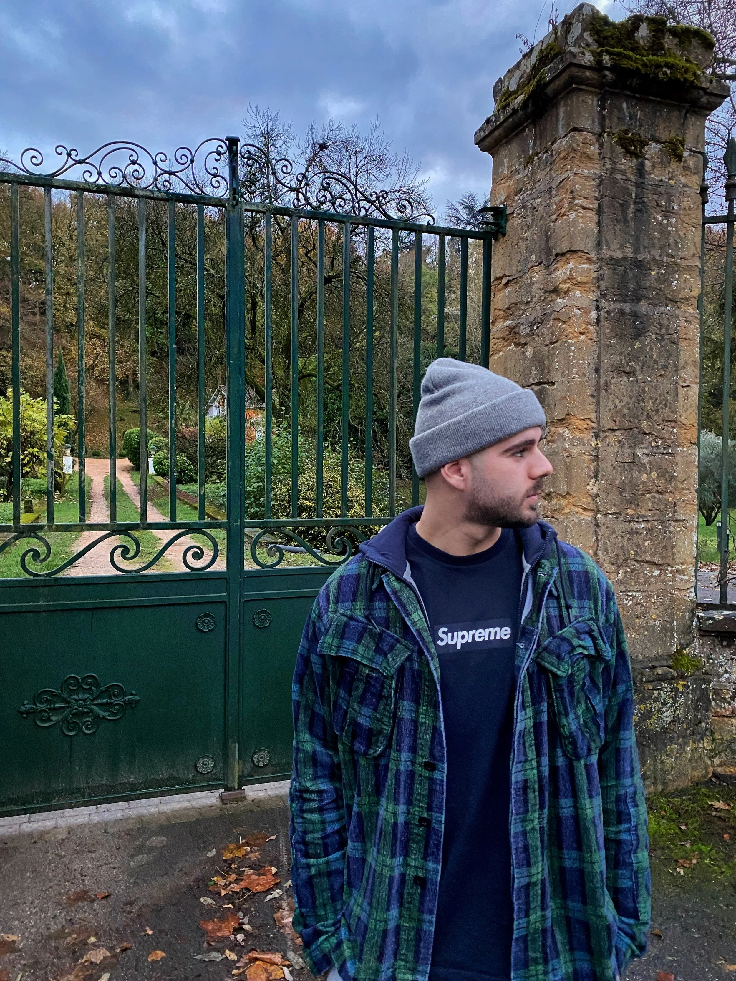 Un jeune homme portant un bonnet gris, un t-shirt Supreme, et une veste à carreaux verts et bleus se tient devant une grille en fer vert et un mur en pierre, dans un jardin avec des arbres et des feuilles mortes.