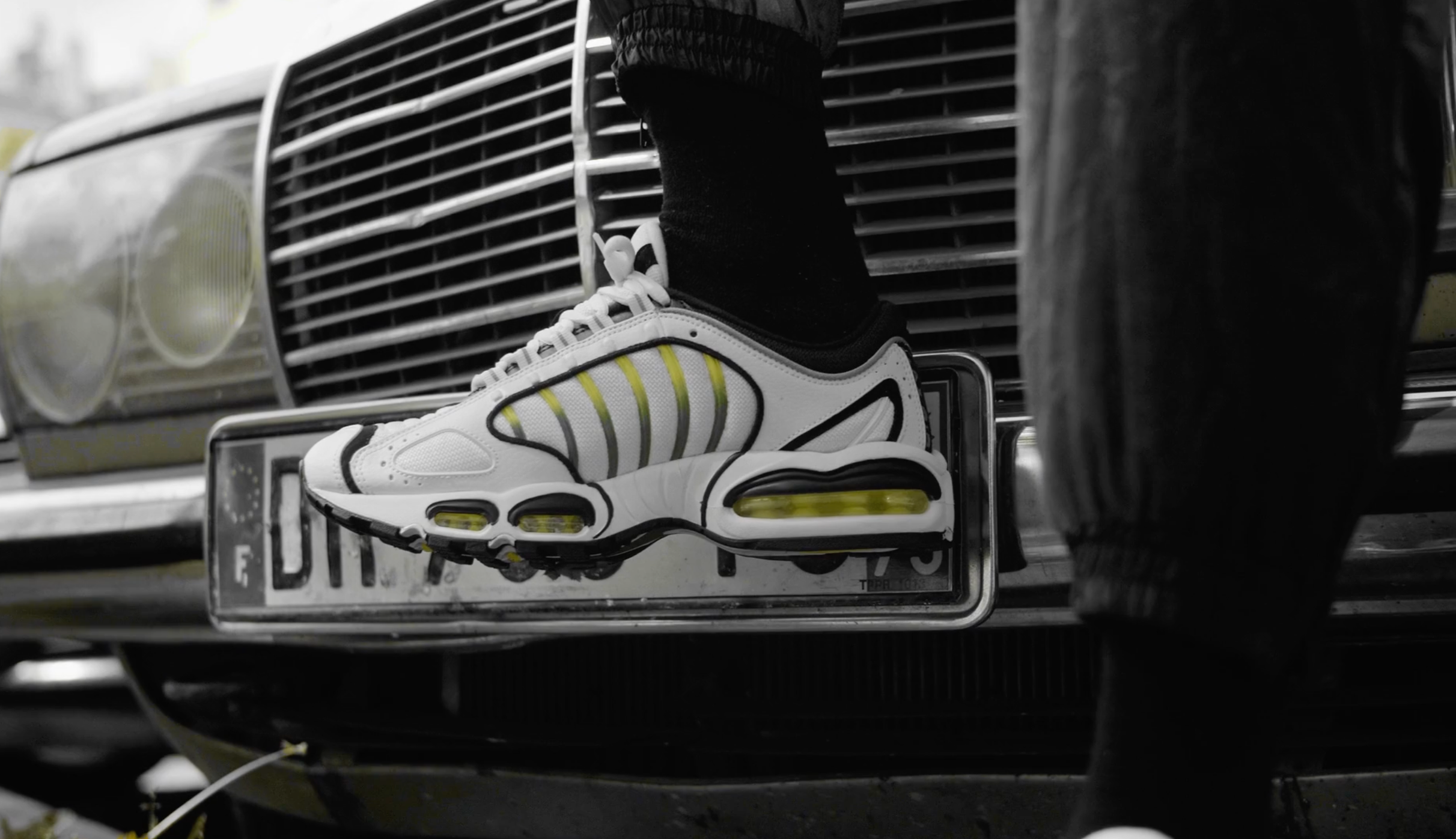 Chaussure de sport blanche avec des accents noirs et jaunes, posée sur une plaque d'immatriculation de voiture, avec une grille de radiateur en arrière-plan.