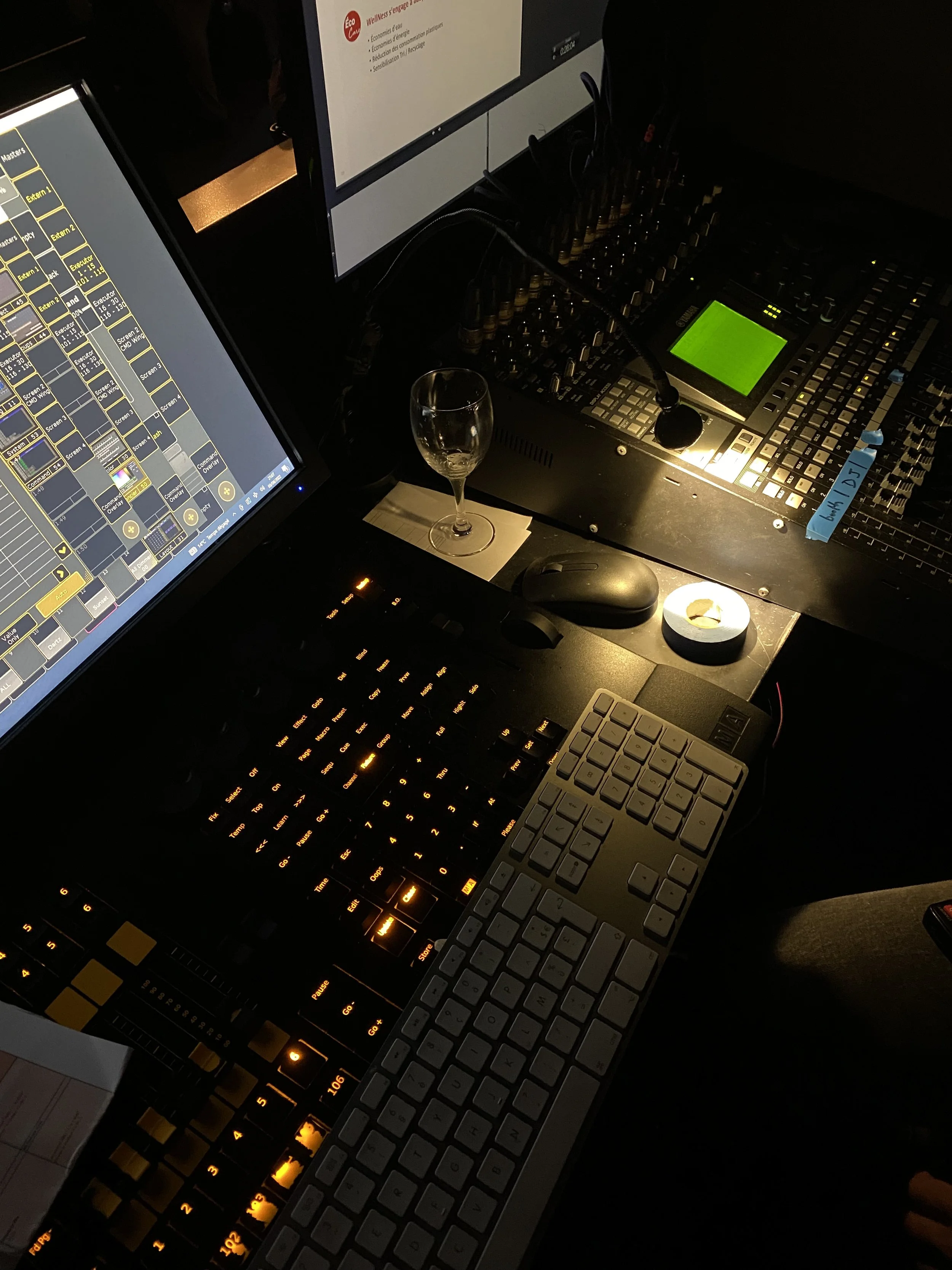 Un poste de travail avec deux écrans d'ordinateur, un clavier, une souris, un microphone, un verre à vin, un snack, et des équipements audio, dans une ambiance sombre.