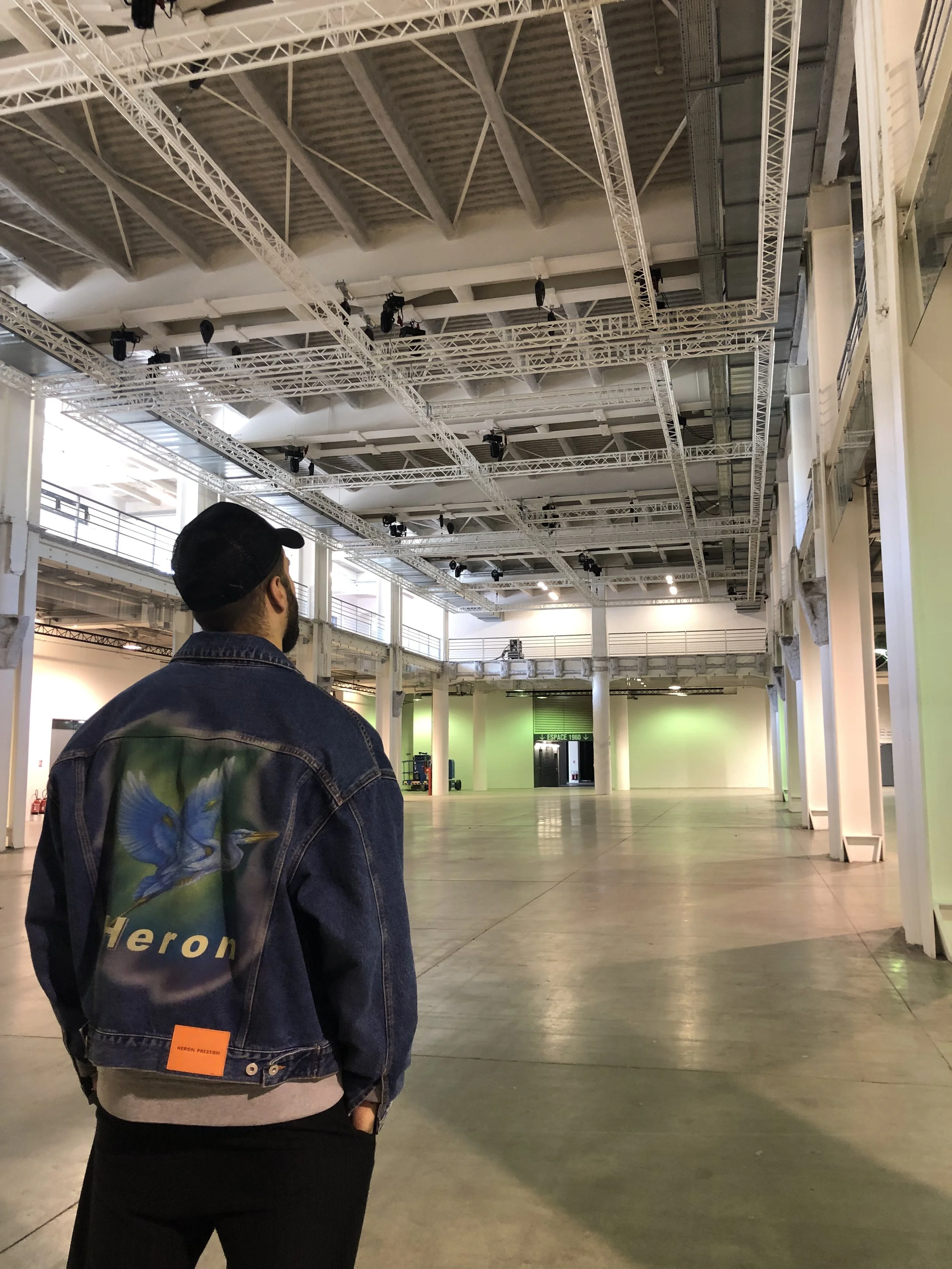 Un homme portant une veste en jean avec un patch d'un héron sur le dos, se tient dans un grand espace intérieur vide avec une structure métallique au plafond et des murs lumineux.