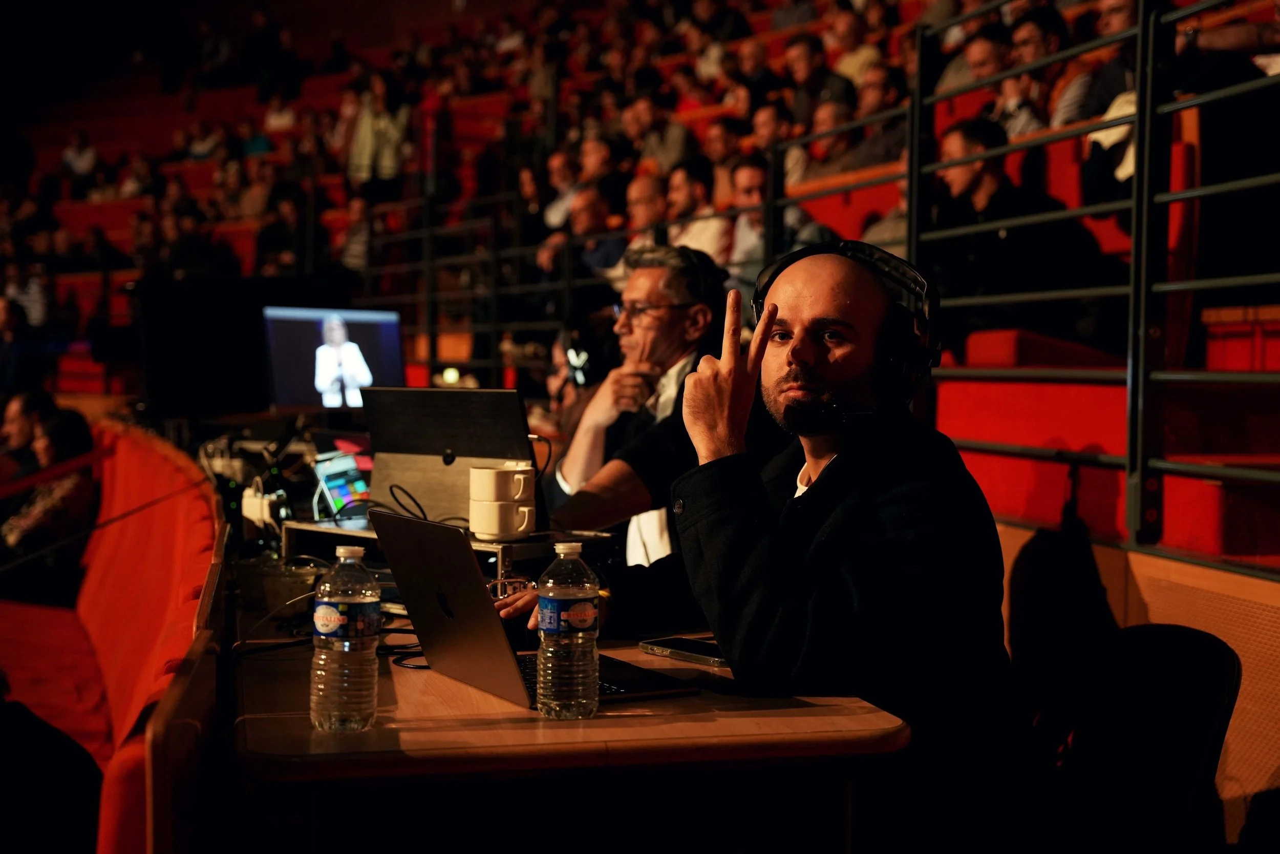 Un homme portant un casque d'écoute et faisant un geste de paix avec ses doigts, assis à une table avec des ordinateurs, dans une salle sombre avec un public en arrière-plan.