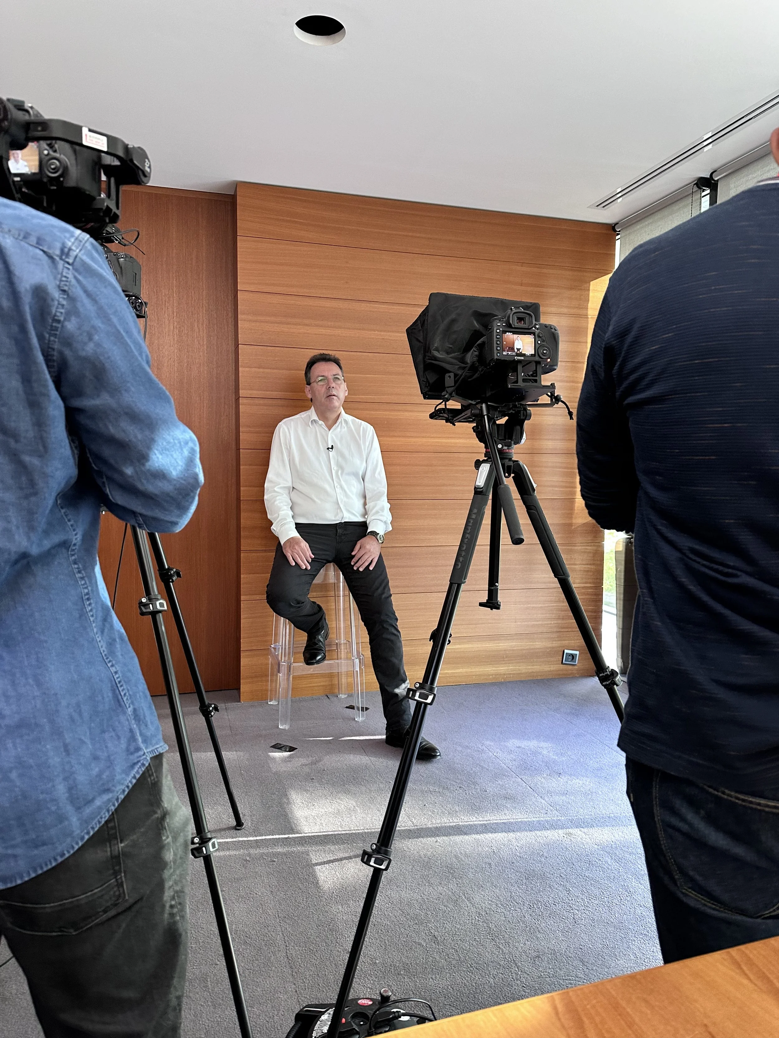 Un homme en chemise blanche assis sur un tabouret de bar, en train de parler lors d'un tournage avec plusieurs caméras et deux hommes en arrière-plan.