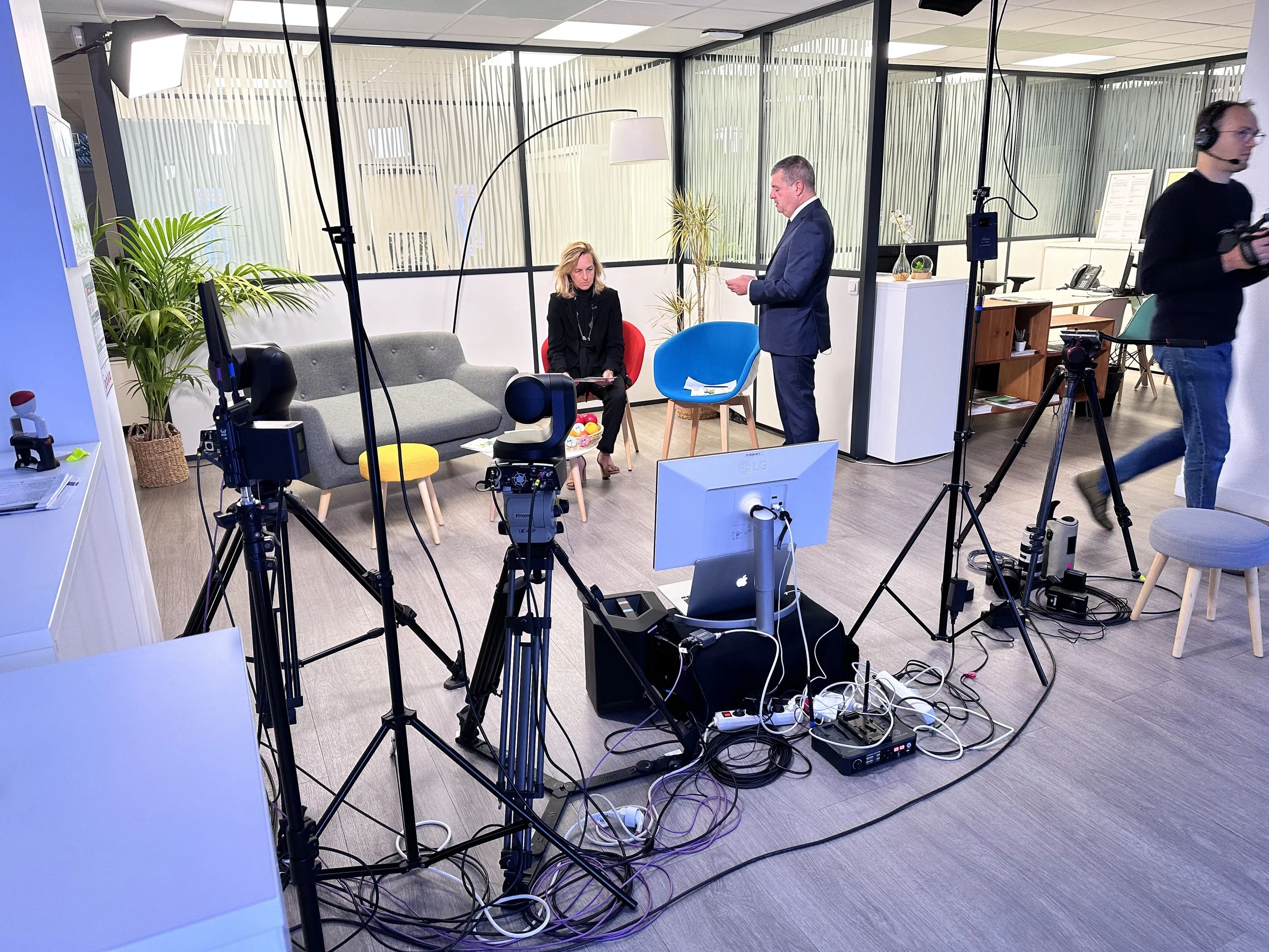 Une salle de réunion ou de studio avec des personnes préparant un enregistrement ou une interview. Il y a des appareils de tournage, des câbles, des lampes, deux personnes assises, une en costume et une other en vêtements noirs, et une autre personne