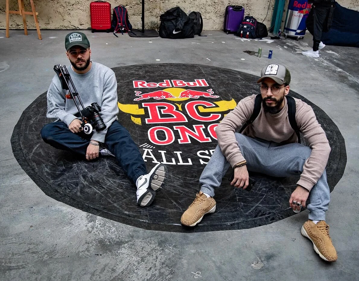 Deux hommes assis sur un logo de Red Bull en forme de cercle sur le sol, à l'intérieur d'un espace au sol en béton, avec des sacs et des valises derrière eux.