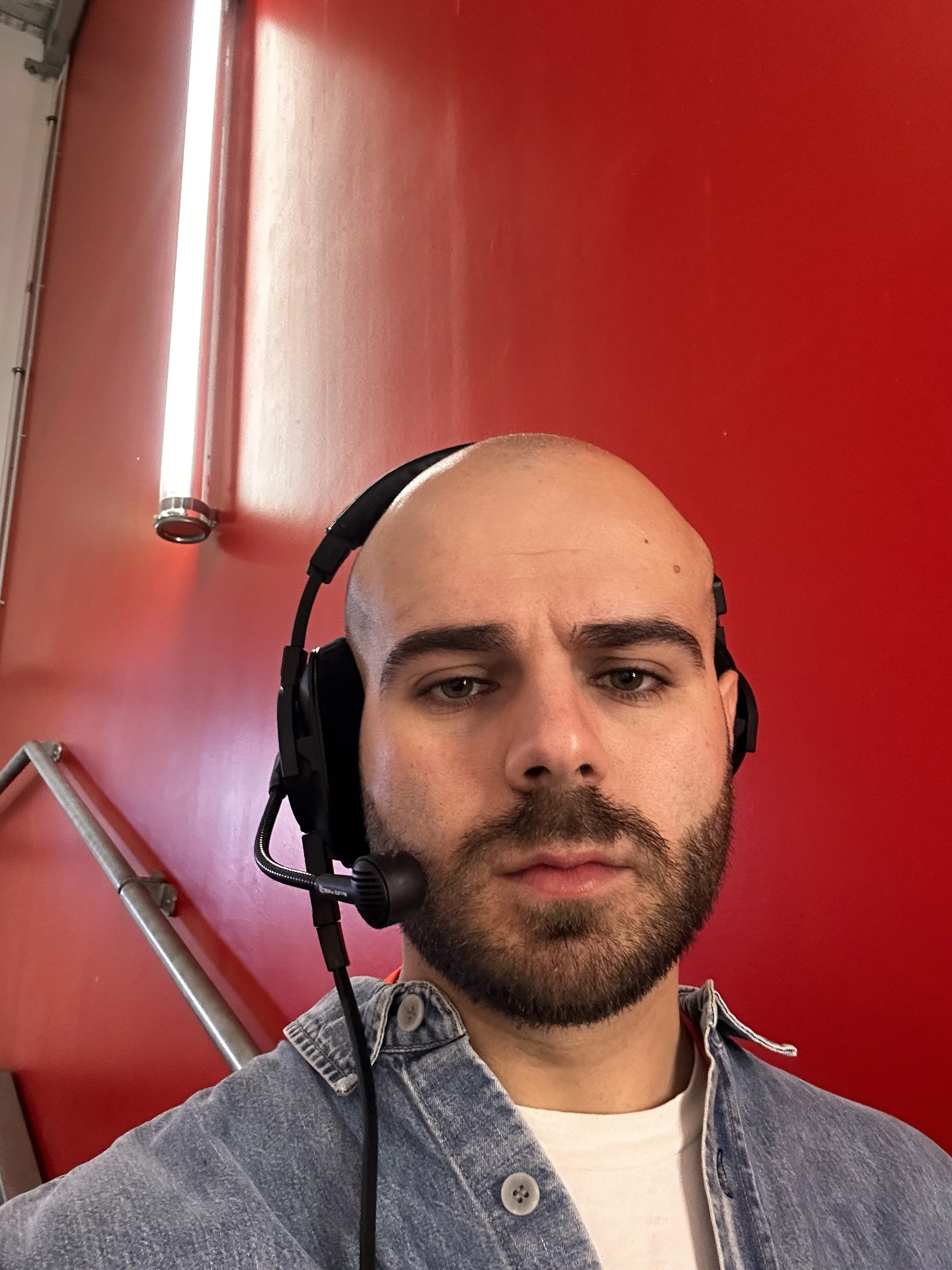 Un homme portant un casque avec microphone, portant une veste en jean, avec un mur rouge en arrière-plan.