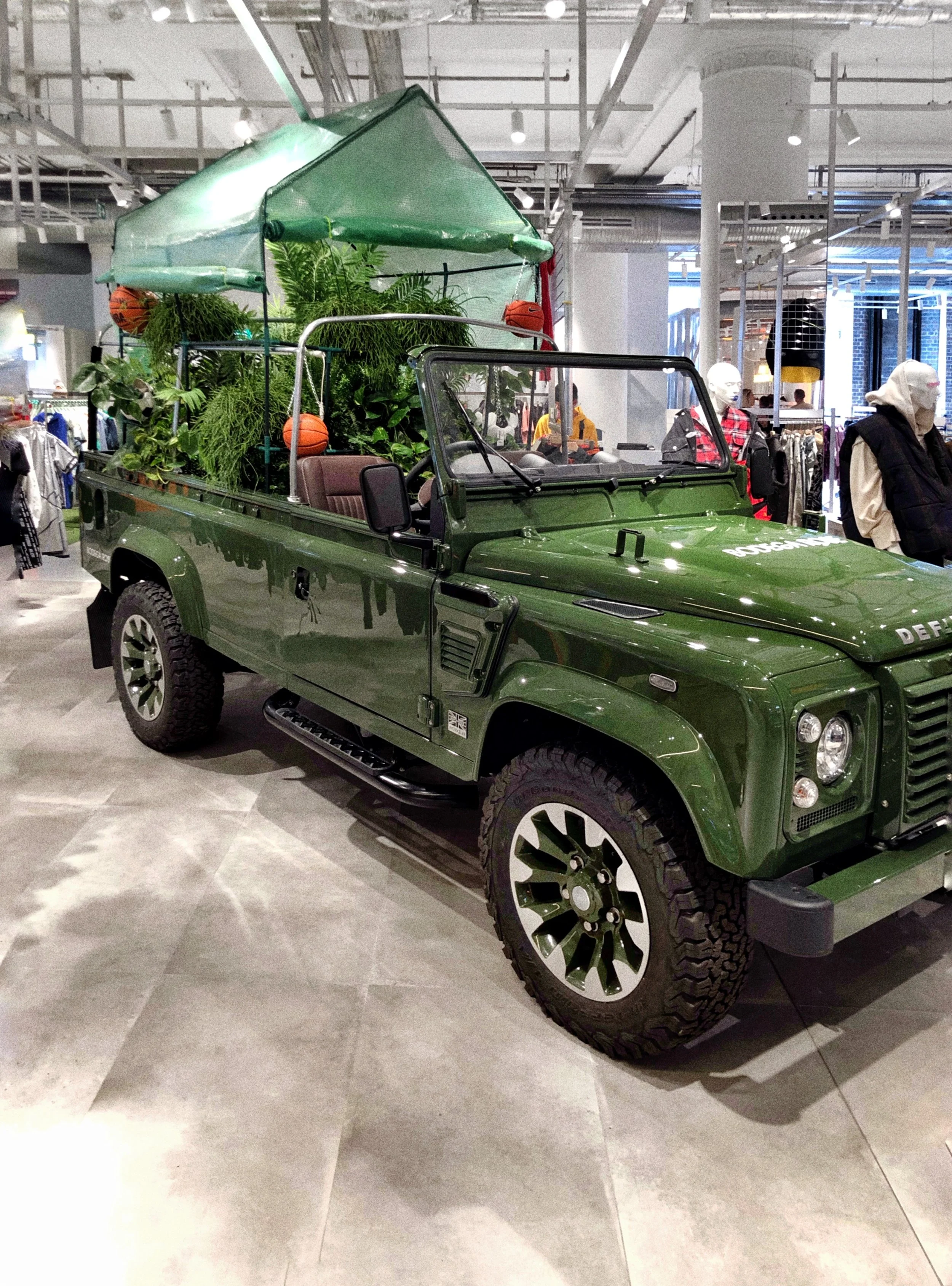 Voiture de chantier Land Rover Defender verte, transformée en un jardin avec des plantes, un parasol vert et des ballons de basket orange, exposée dans un magasin.