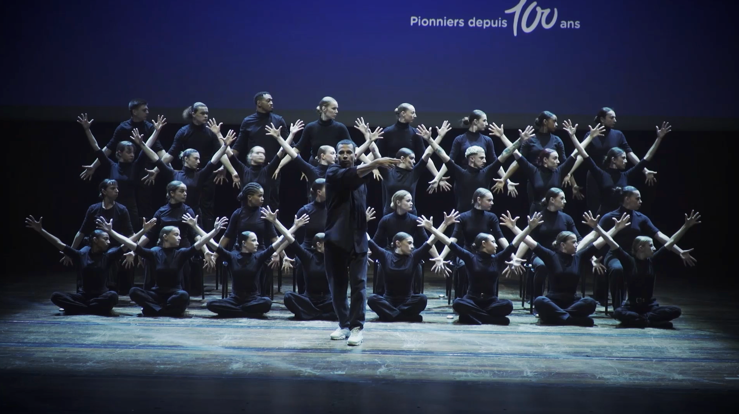 Groupe de danseurs assis et debout en position de performance sur scène, vêtus de noir, avec un homme au centre qui semble diriger. Un écran en arrière-plan affiche le texte "Pionniers depuis 100 ans".