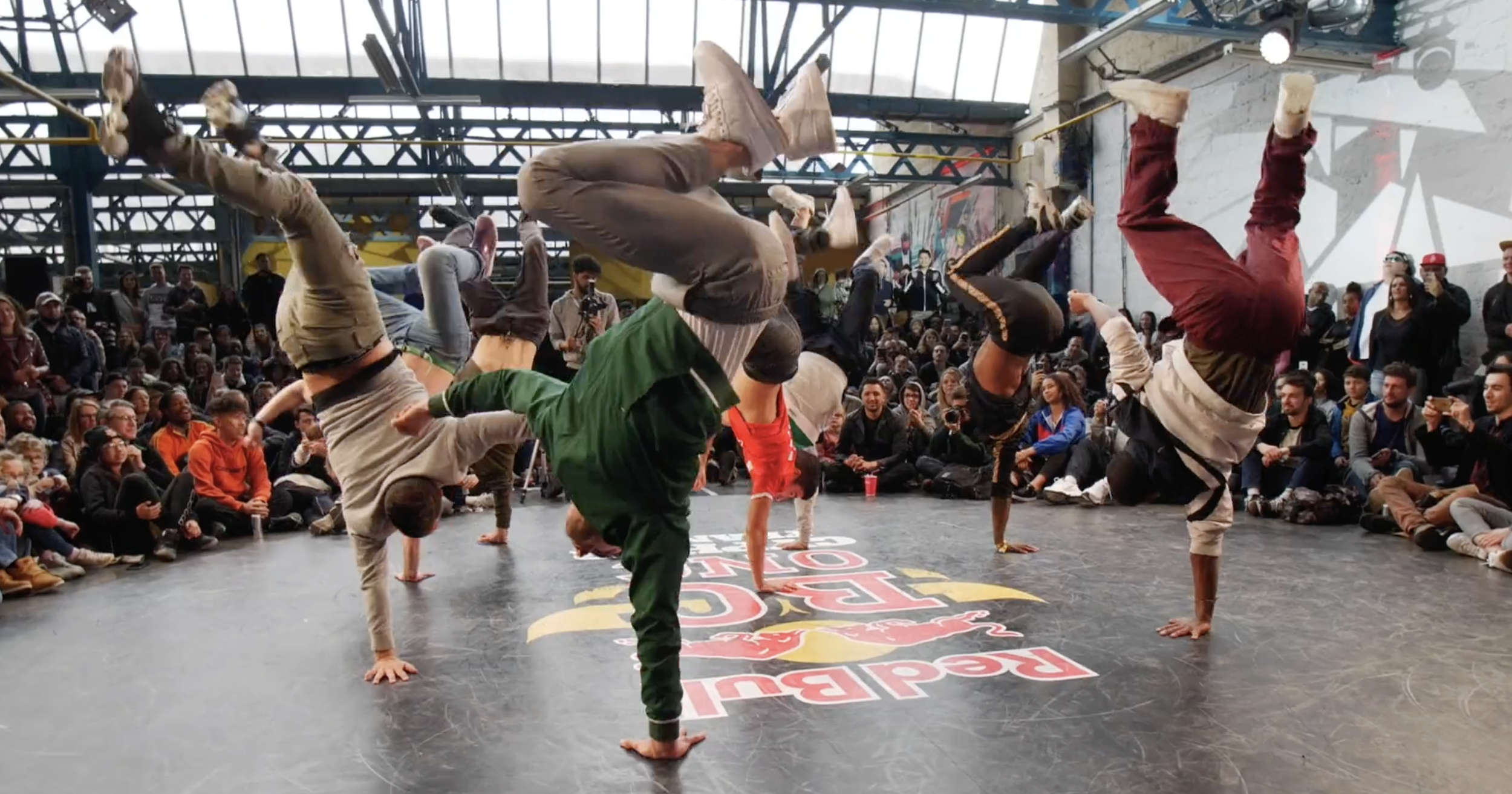 Des danseurs de breakdance exécutant des figures en équilibre sur leurs mains lors d'une compétition ou démonstration, entourés d'un public assis et debout dans un espace industriel.