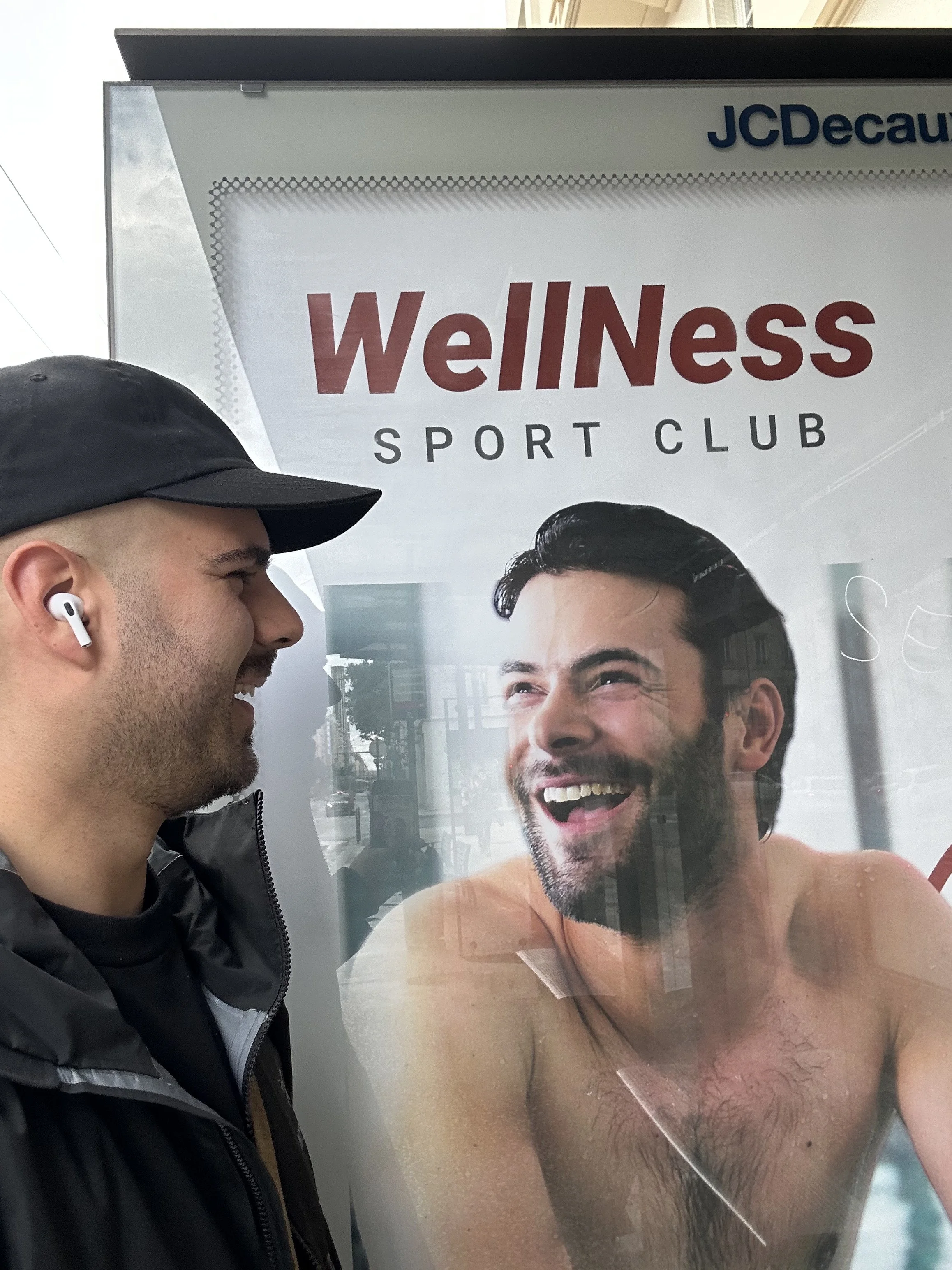 Un homme souriant et portant des écouteurs regarde une affiche de spa ou de centre de bien-être avec un homme nu dans une piscine ou un spa.