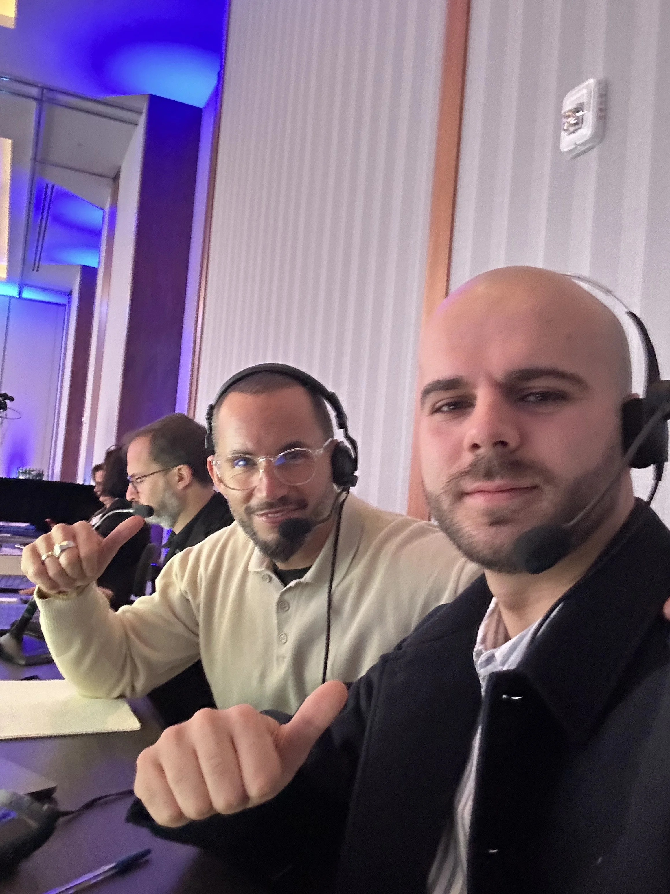 Trois hommes assis à une table, portant des casques avec microphones, dans une salle avec un éclairage bleu. Deux d'entre eux montrent le signe du pouce en l'air.