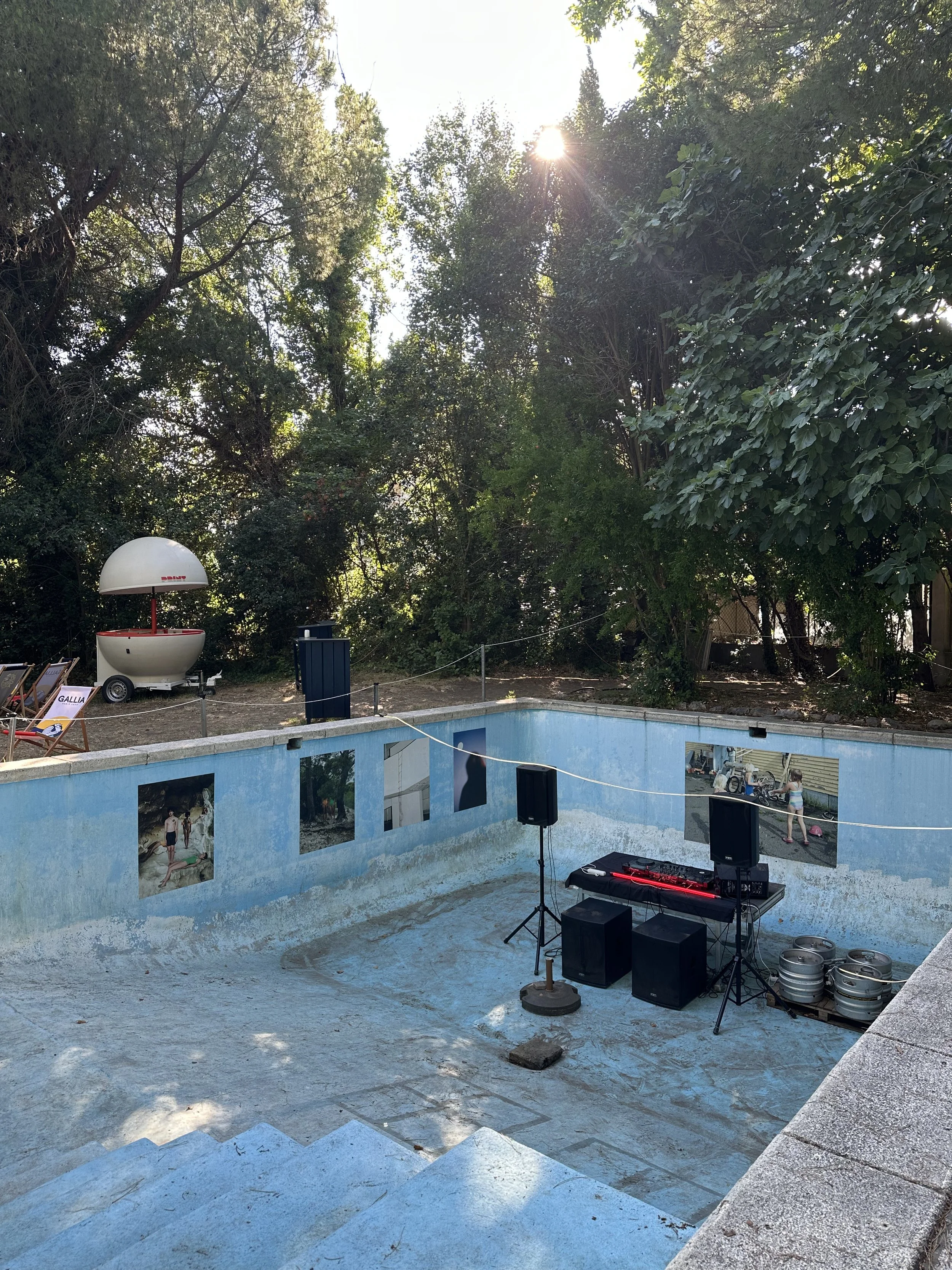 Piscine abandonnée transformée en espace pour un événement ou une fête, avec une table de mixage, haut-parleurs, et des affiches sur le mur. Sur le côté, un siège de plage avec une affiche 'Gallia'. Des arbres et le soleil en arrière-plan.