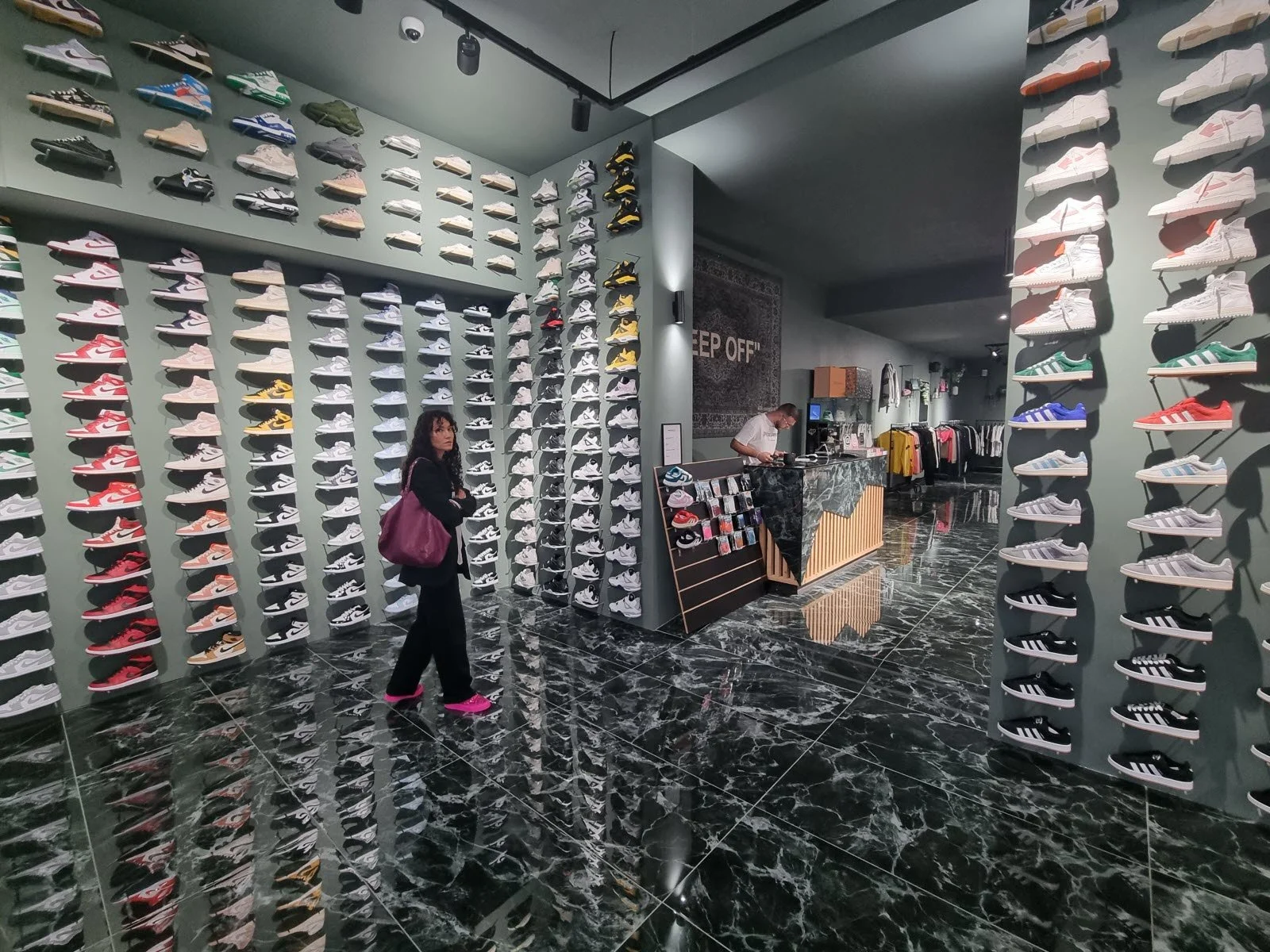 Interior d'une boutique de sneakers avec murs remplis de chaussures de différentes couleurs et modèles, un comptoir de caisse et un client portant un sac violet.
