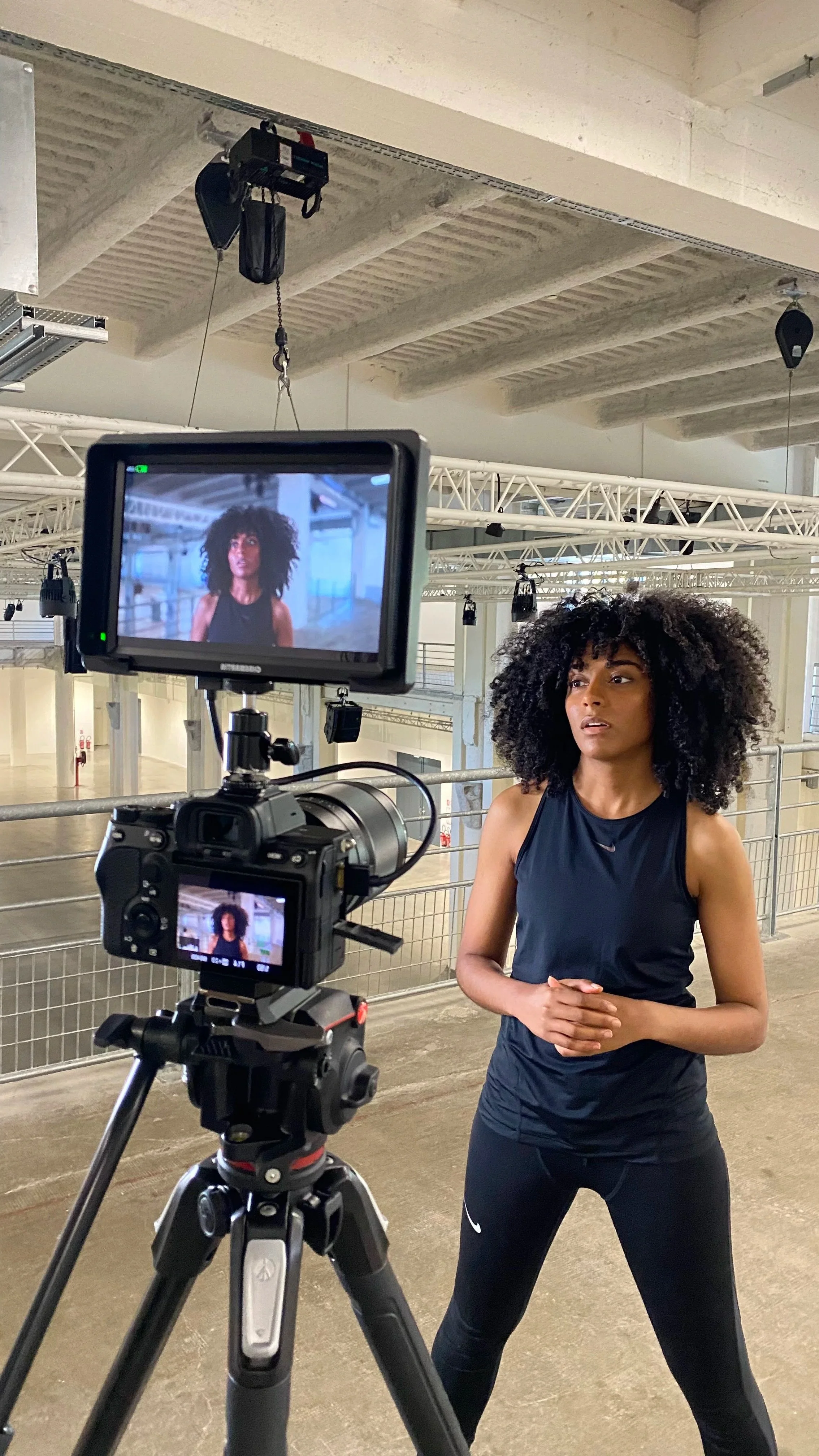 Une femme avec des cheveux frisés noirs longs, portant un débardeur noir et un pantalon noir, en train de parler à la caméra dans ce qui semble être un studio ou un gymnase, lors d'une séance d'enregistrement ou de tournage.