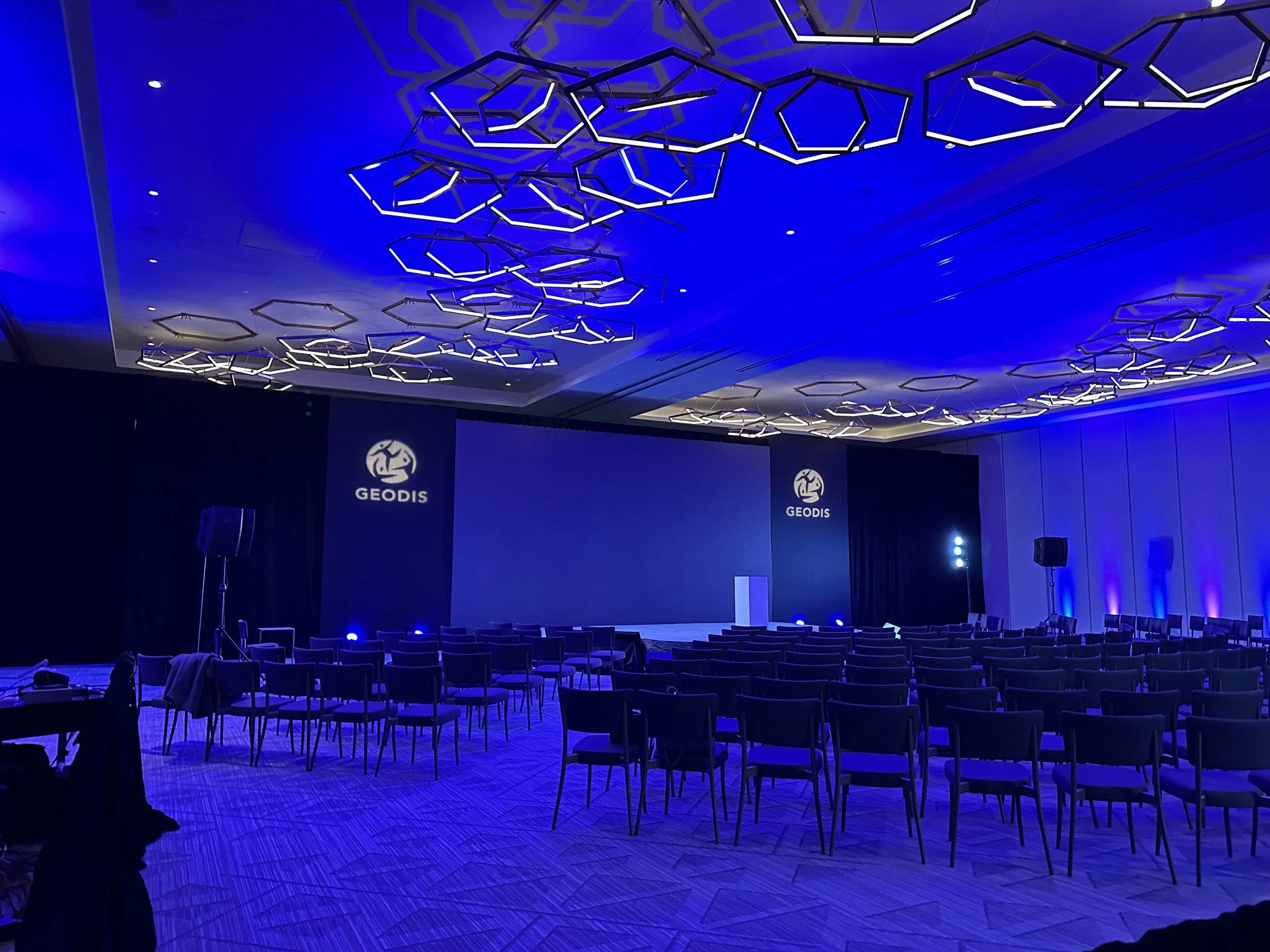 Salle de conférence avec plafond orné de luminaires en forme d'hexagones. La scène principale a un grand écran et deux panneaux avec le logo GEODIS. Sièges alignés pour un auditoire, éclairage tamisé avec une tonalité bleue.