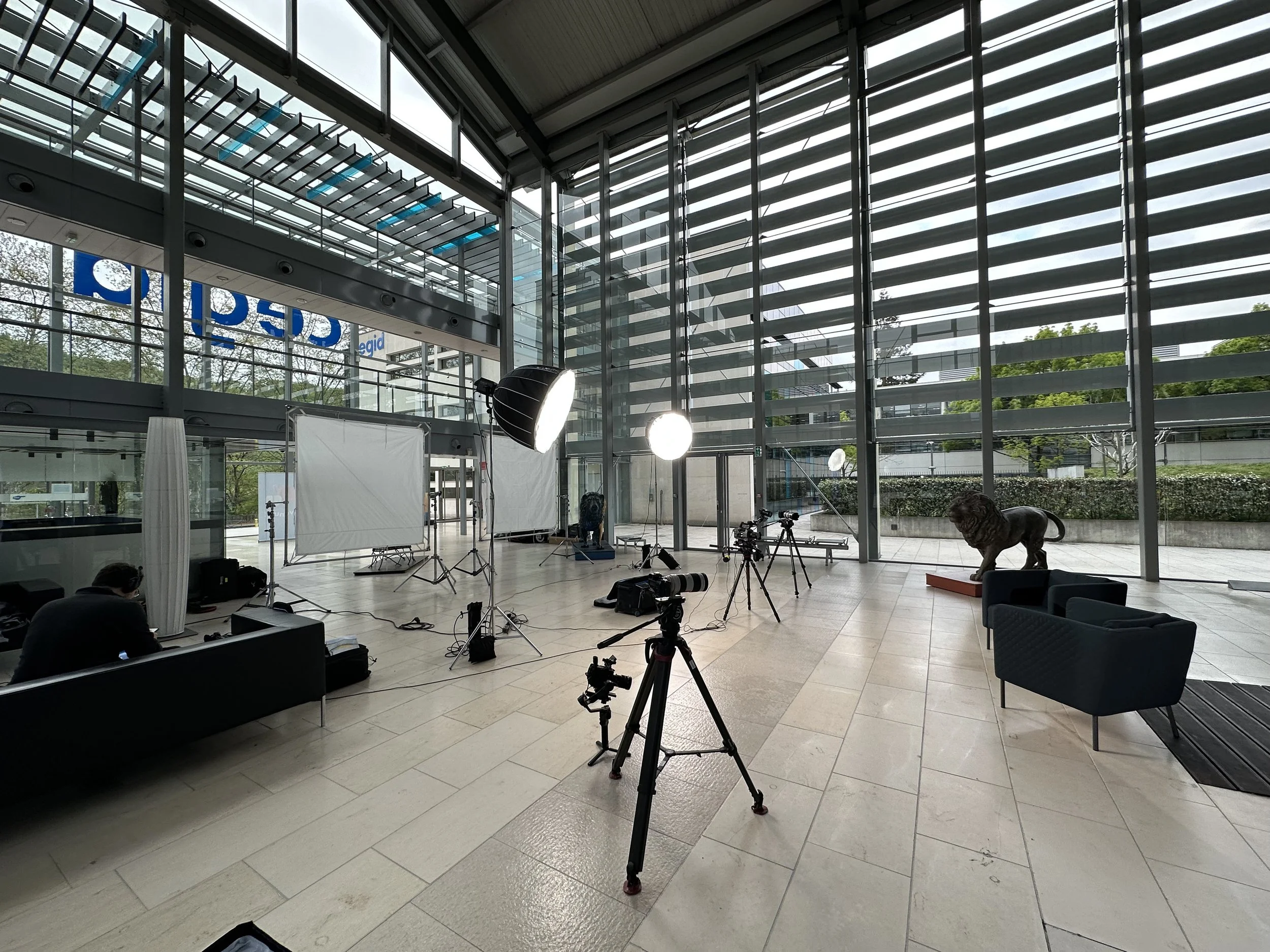 Un studio de tournage intérieur avec équipements de photographie, deux écrans, des lampes, des trépieds, des sièges, une statue de lion et de larges fenêtres en verre.