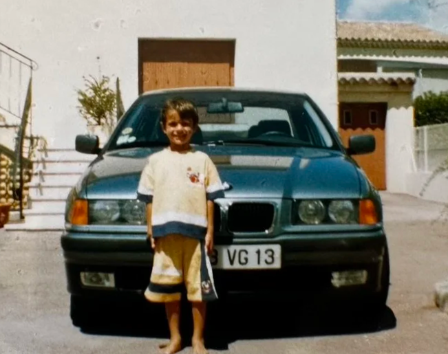 Enfant souriant debout devant une voiture bleue, dans une cour résidentielle avec une maison en arrière-plan.