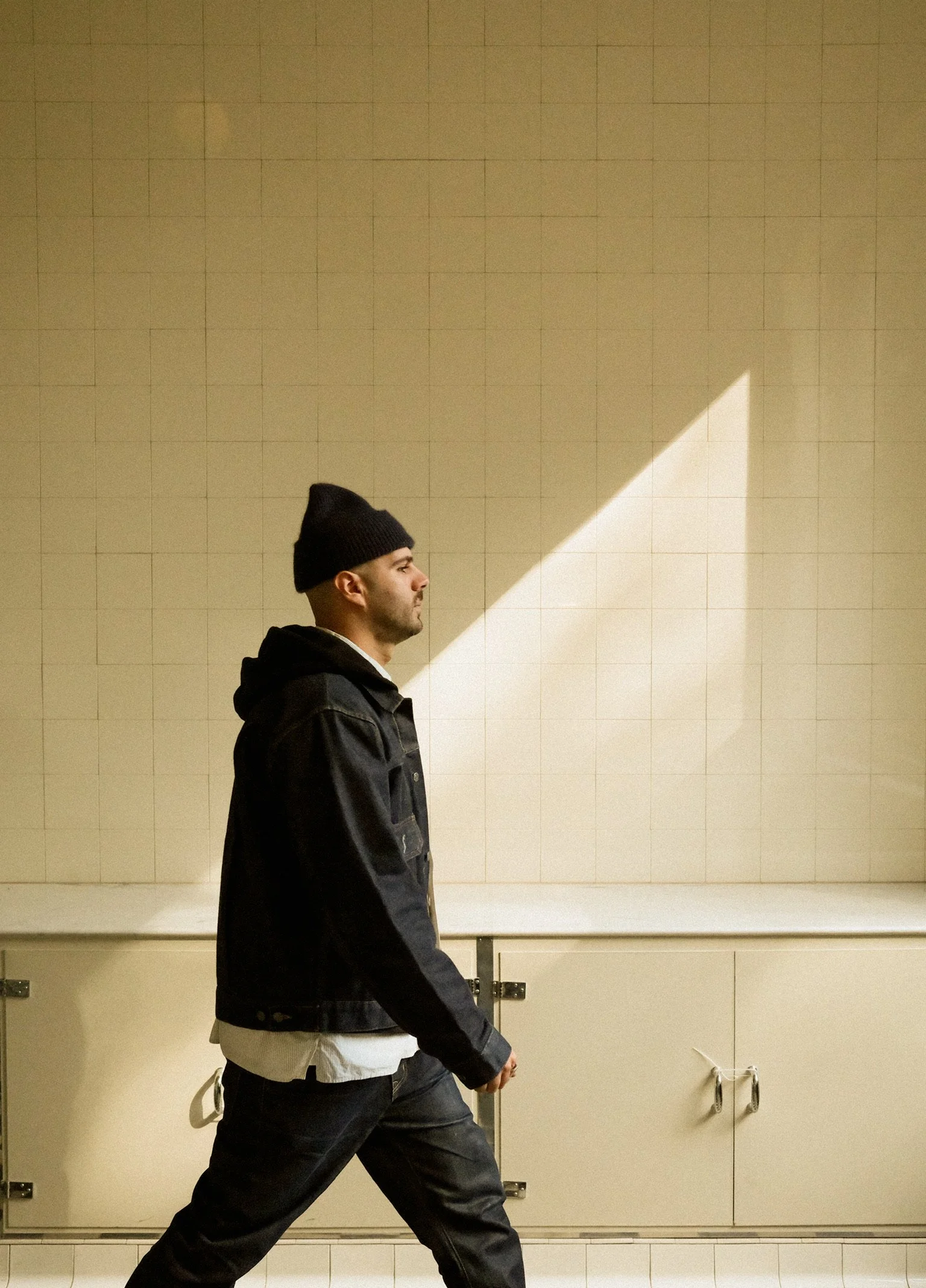 Un homme marche dans une pièce, portant un bonnet noir, une veste en jean noire, et une chemise blanche. La lumière crée un contraste avec un éclairage diagonal sur le mur beige en arrière-plan.