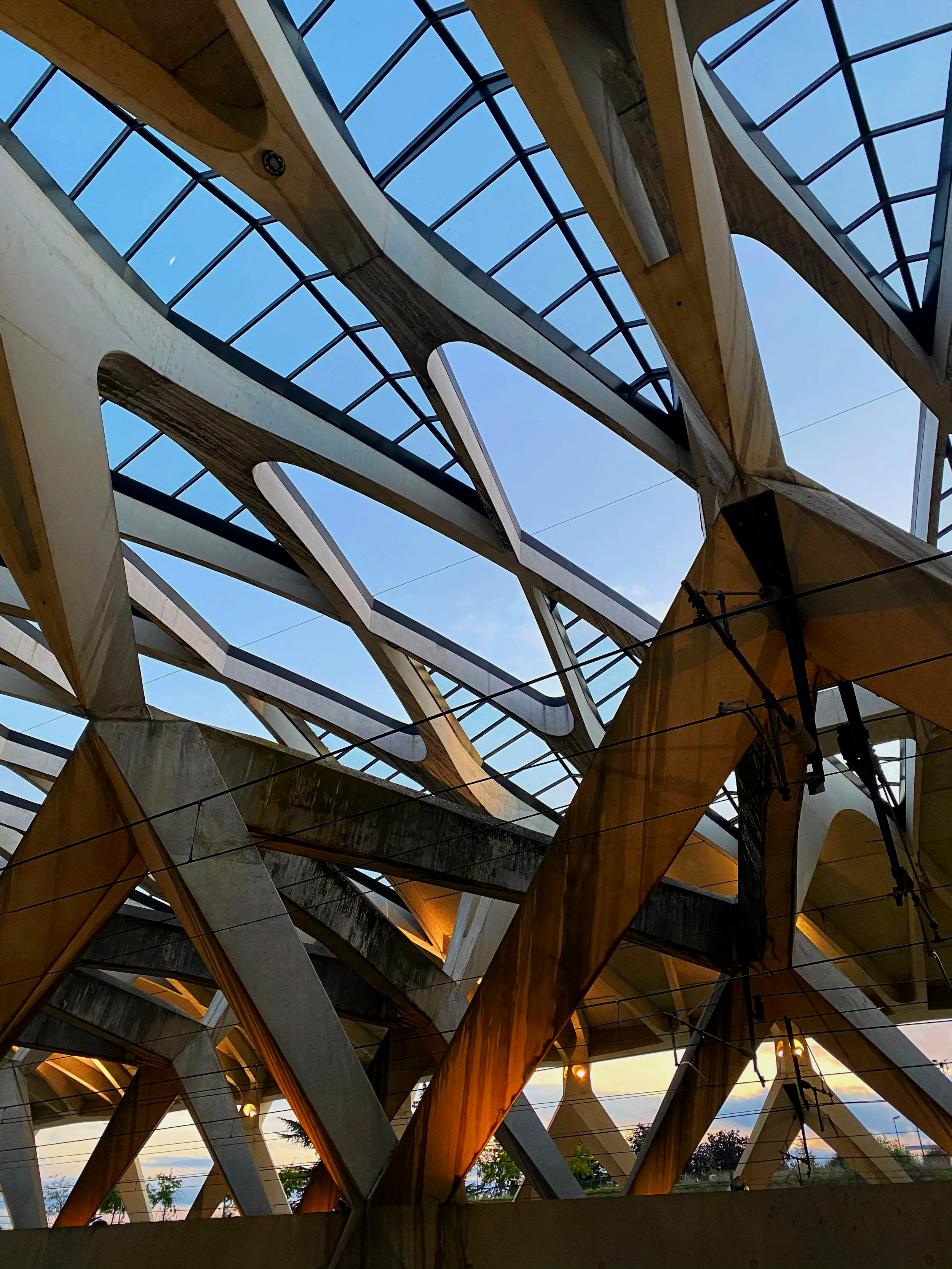 Une structure architecturale moderne faite de bois, de béton et de verre, vue de l'intérieur, accentuant des formes angulaires et courbes avec un ciel bleu en arrière-plan.