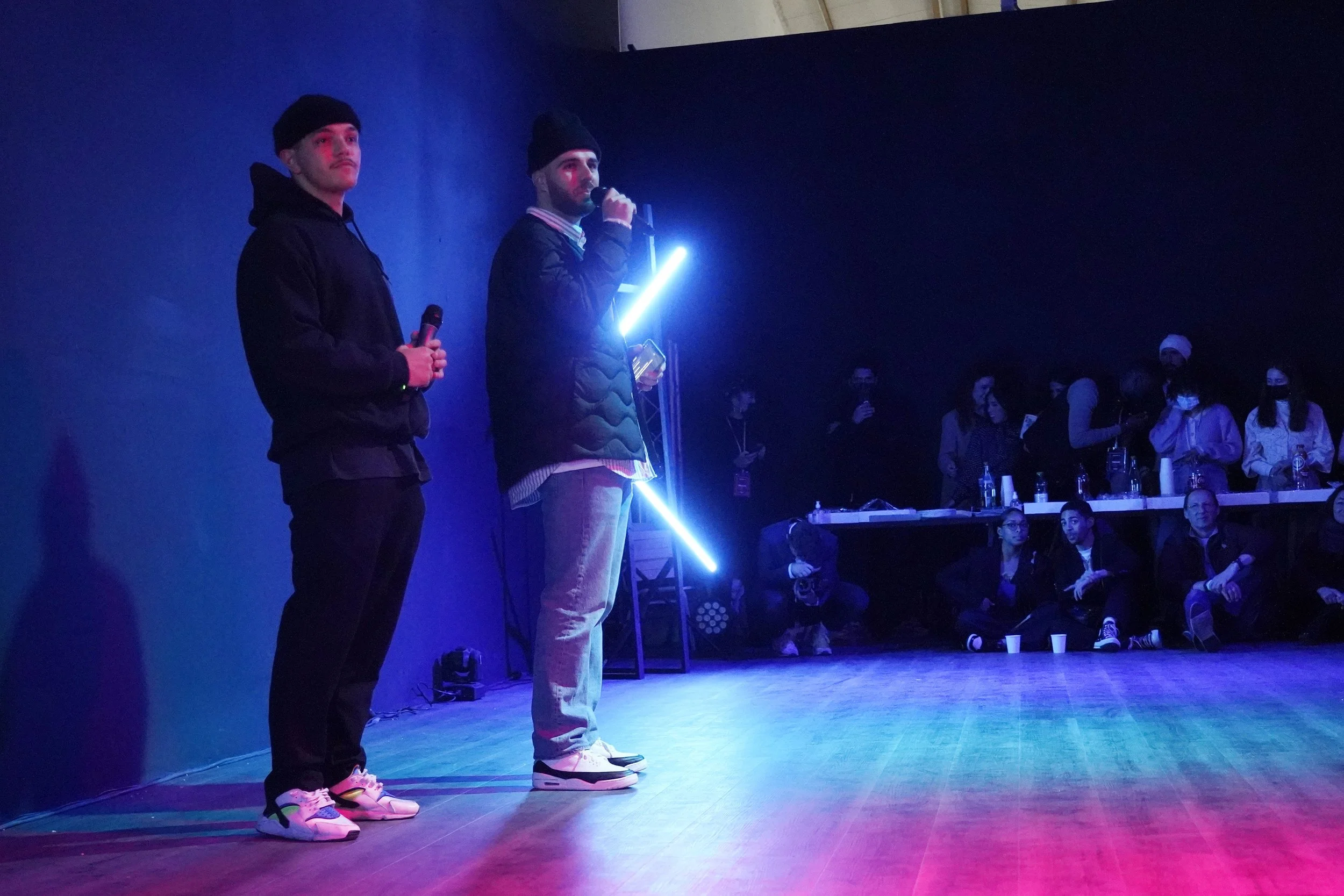 Deux jeunes hommes portant des casquettes noires, l'un en hoodie noir et l'autre en veste, tenant des microphones sur une scène avec un fond sombre, dans un environnement lumineux avec des effets colorés. Plusieurs personnes sont assises ou debouts à