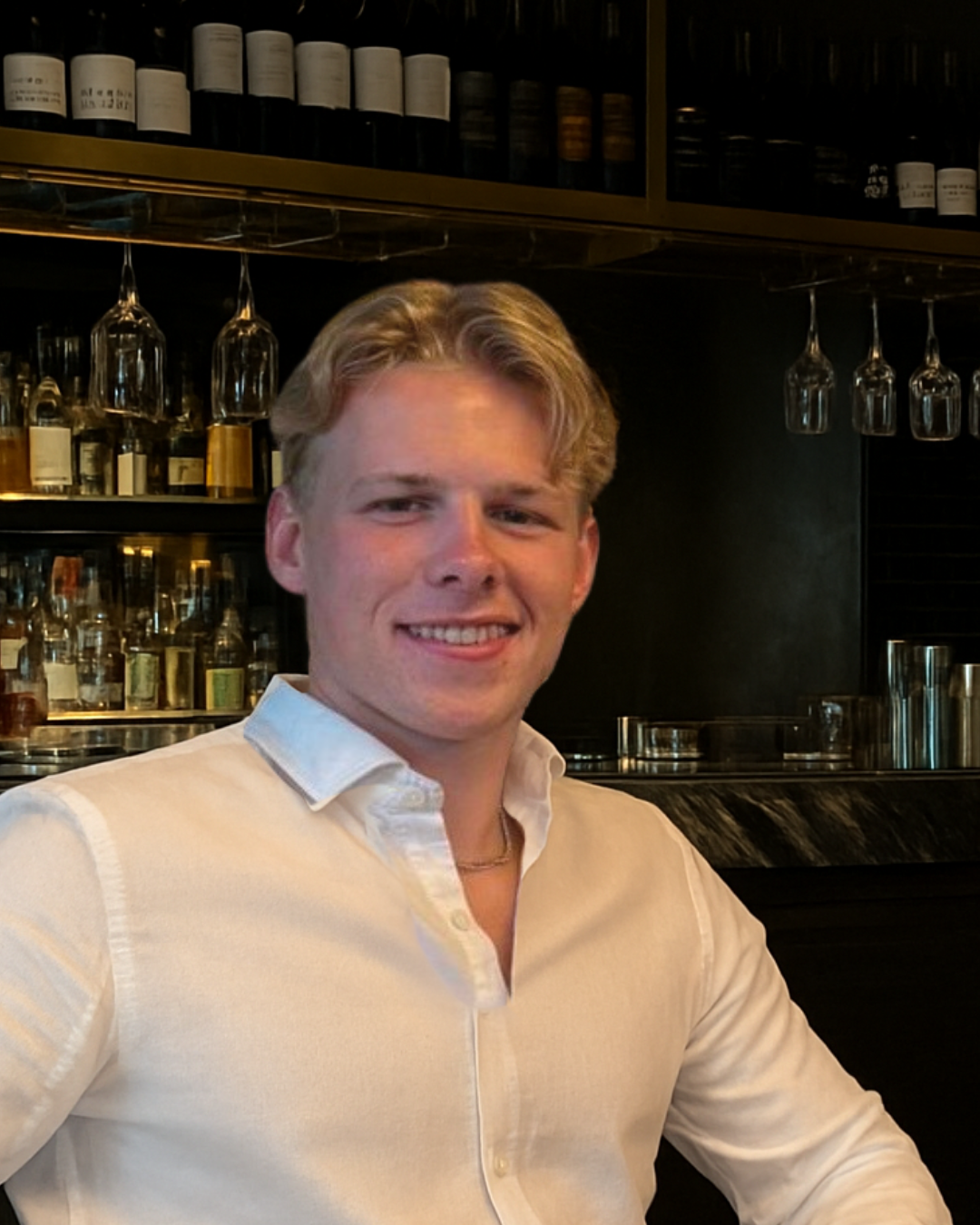 A young man with blond hair smiling at the camera, wearing a white shirt, sitting in a bar or restaurant with shelves of bottles and hanging glasses in the background.