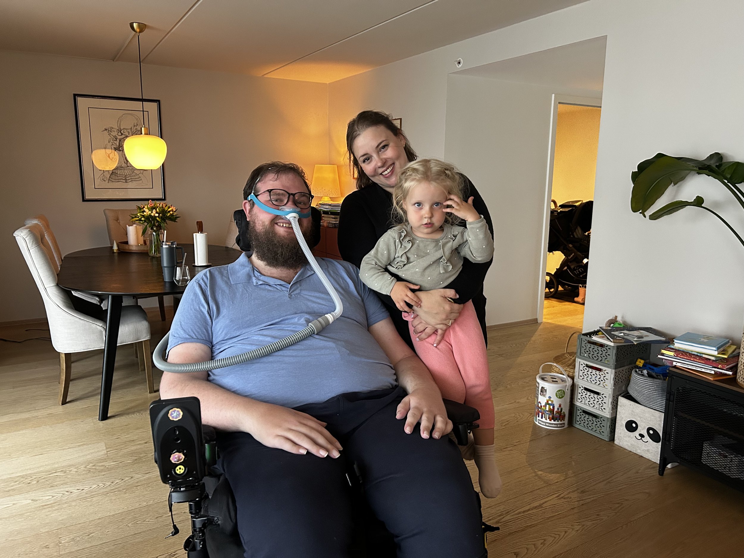 Jostein Altøy Opsahl sitter i en elektrisk rullestol med pustemaskin og smiler mot kamera. Inntil han står samboeren Heidi Hammervold Hauge og smiler mot kamera med datteren Thekla på armen. Fotografi.